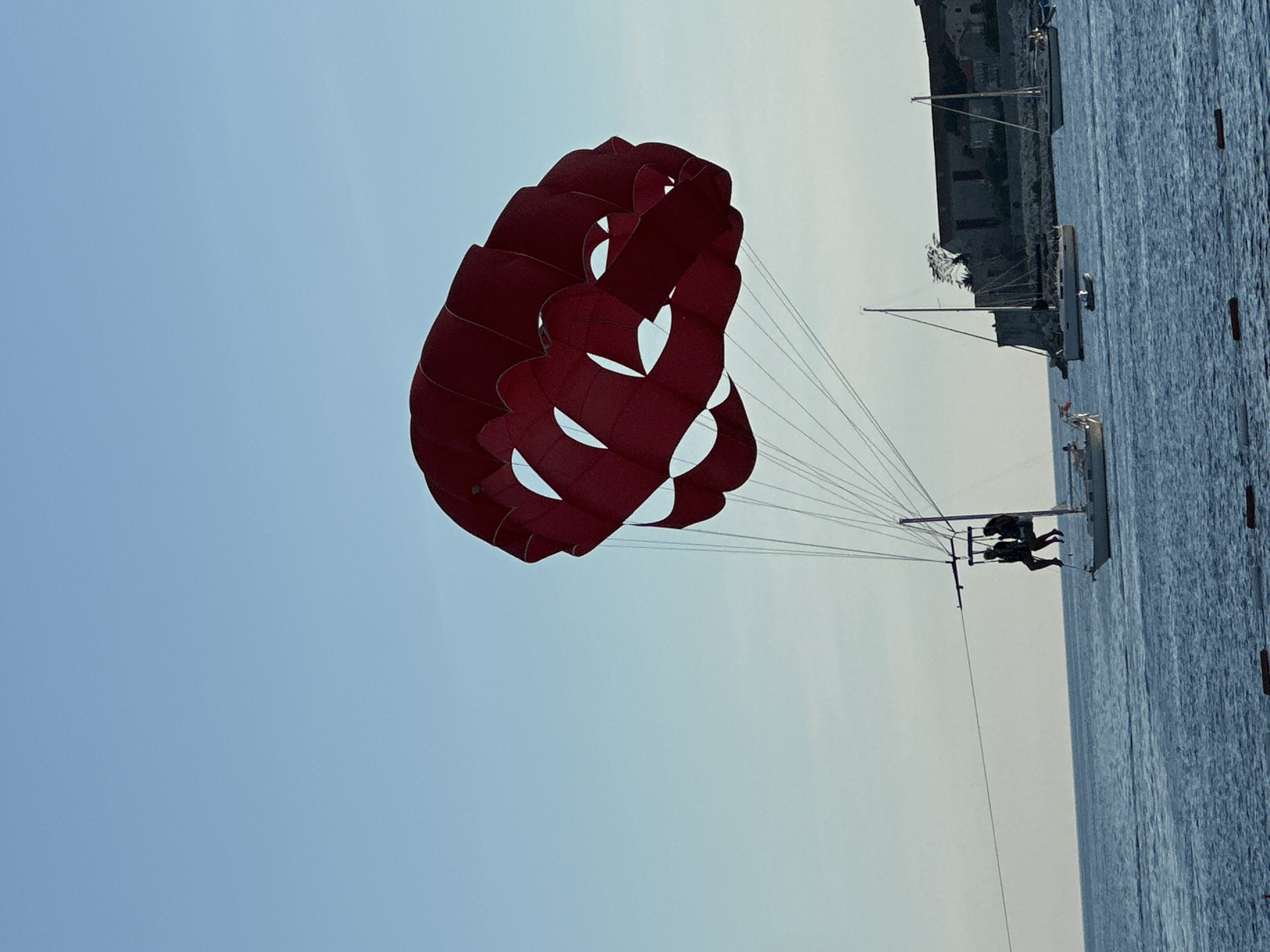 In the foreground, a large, deep red parasail dominates the frame against a clear, pale blue sky. Suspended below the parasail by thin white lines are two silhouetted figures, appearing to be seated side-by-side, attached to a harness system. One figure appears slightly taller and positioned behind the other. To the right and slightly behind them, a sailboat with a tall mast and white hull is visible, with a shorter, dark-colored dinghy tethered to its stern. The sailboat is in motion, its sails furled.
In the mid-ground, several other sailboats are anchored or drifting on the calm, deep blue sea. One sailboat with a single mast and white hull is prominently positioned in the center of the image. Another sailboat, further to the right, also features a single mast and white hull. A small, dark dinghy is situated between these two sailboats.
In the background, a fortified stone structure, possibly an old castle or fortress, is built into a hillside overlooking the water. The stone walls are weathered and appear to be made of rough-hewn blocks. Several buildings with red-tiled roofs are visible within the fortified area. Trees are sparsely scattered on the hillside. The overall scene suggests a coastal location with recreational activities taking place on the water, likely during the daytime. Given the architecture of the fortress and the coastal setting, this scene is identifiable as Budva, Montenegro.