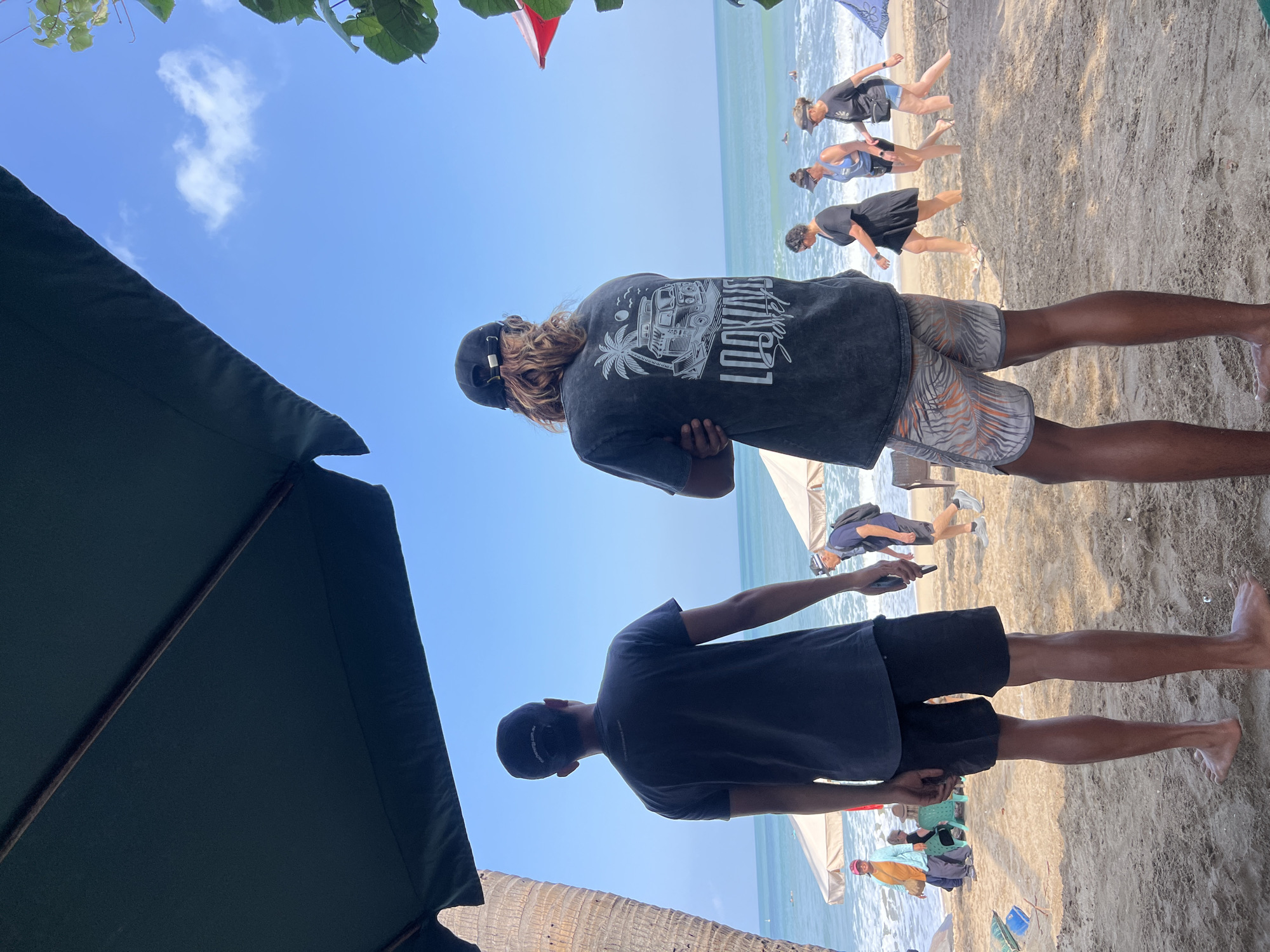 Two men stand on a sandy beach in Kuta, Indonesia, facing away from the camera towards the ocean. The man on the left wears a dark t-shirt and black shorts, while the man on the right wears a dark t-shirt with a graphic print and patterned shorts. In the background, several people walk along the shoreline, and beach umbrellas are visible. The sky is clear blue with a few scattered clouds. The ocean is calm, with gentle waves reaching the shore. Palm trees and foliage frame the edges of the scene. The overall atmosphere is relaxed and summery.