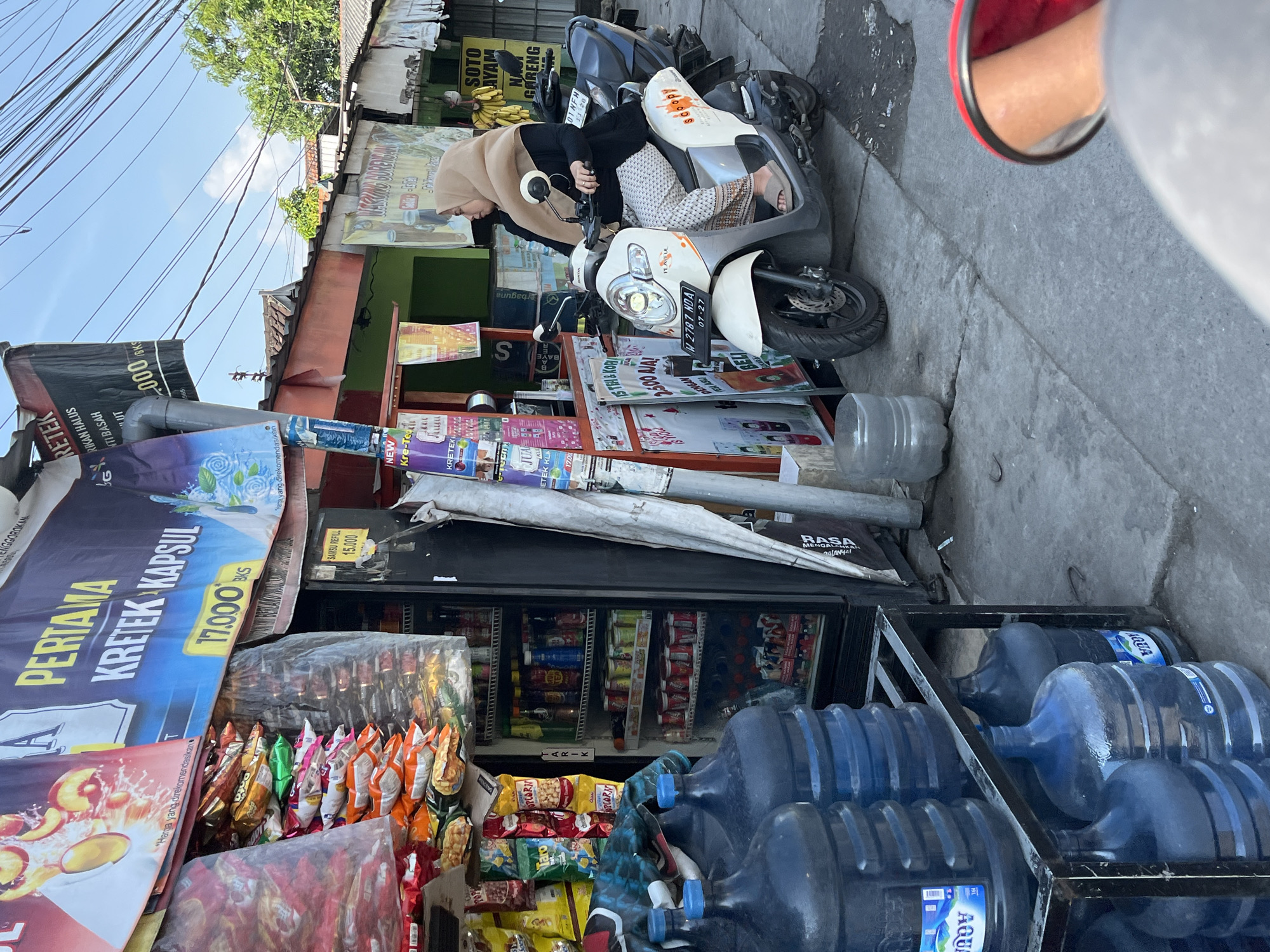In Tulangan, Indonesia, a street-level view captures a modest storefront offering various goods, including bottled water, snacks, and what appears to be tobacco products, indicated by signage featuring "Kretek." A woman wearing a hijab and patterned pants sits on a white scooter parked beside the store. She is looking to the right, away from the camera. The store itself is stocked with products visible through a glass-fronted refrigerator and on shelves outside. A banner above the store displays the text "PERTAMA KRETEK KAPSUL" and a price of "17.000 BKS." The environment is consistent with a street scene in Indonesia, with overhead wires, tropical trees, and paved ground. The lighting suggests it is daytime.