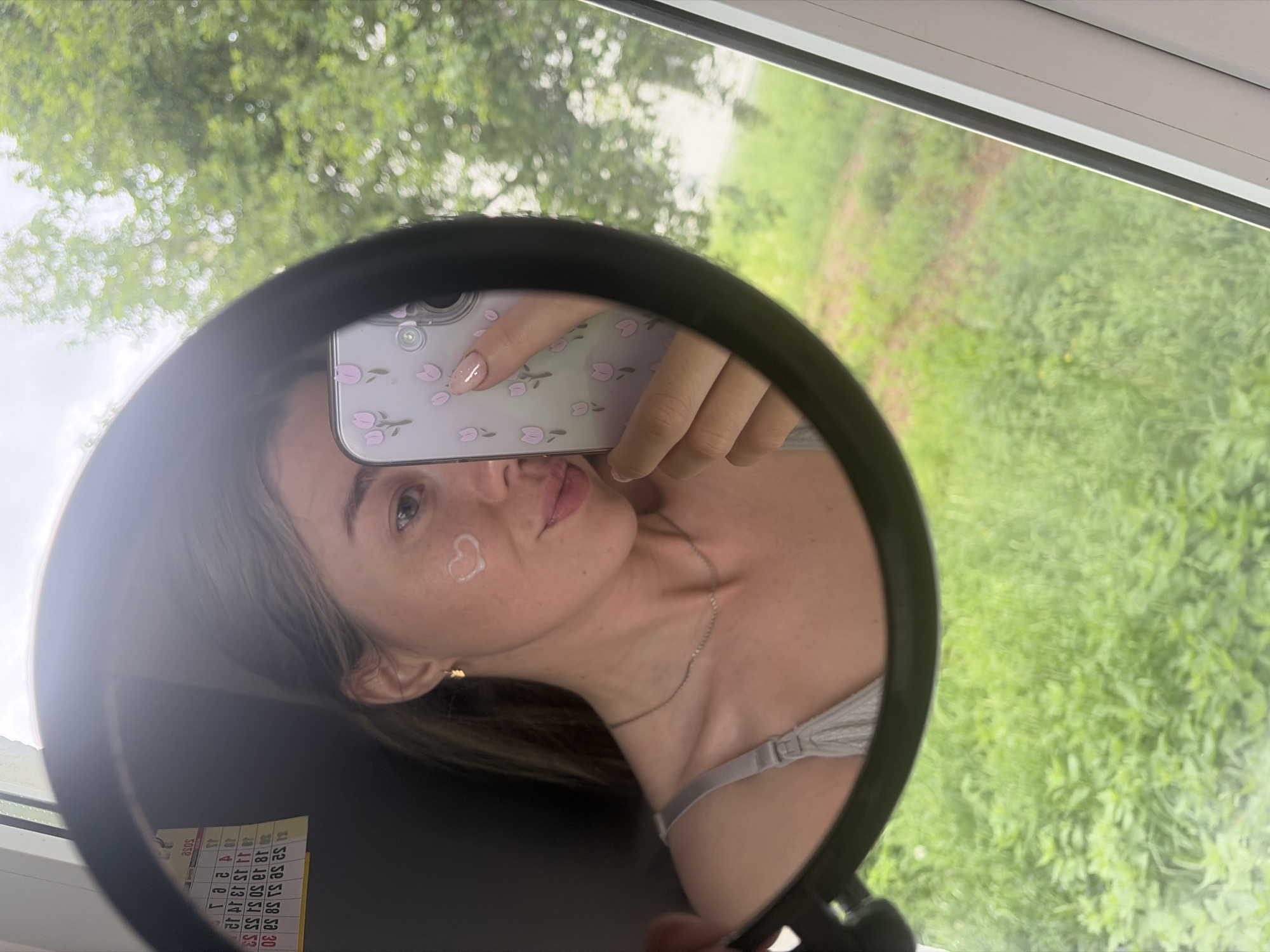 The image shows a young woman reflected in a round mirror, holding a smartphone with a floral case to take a selfie. She has a small heart-shaped design on her cheek. She wears a thin silver necklace. Behind the mirror, there's a view of green foliage, possibly trees and grass. The location appears to be Chaikovsky, Russia.