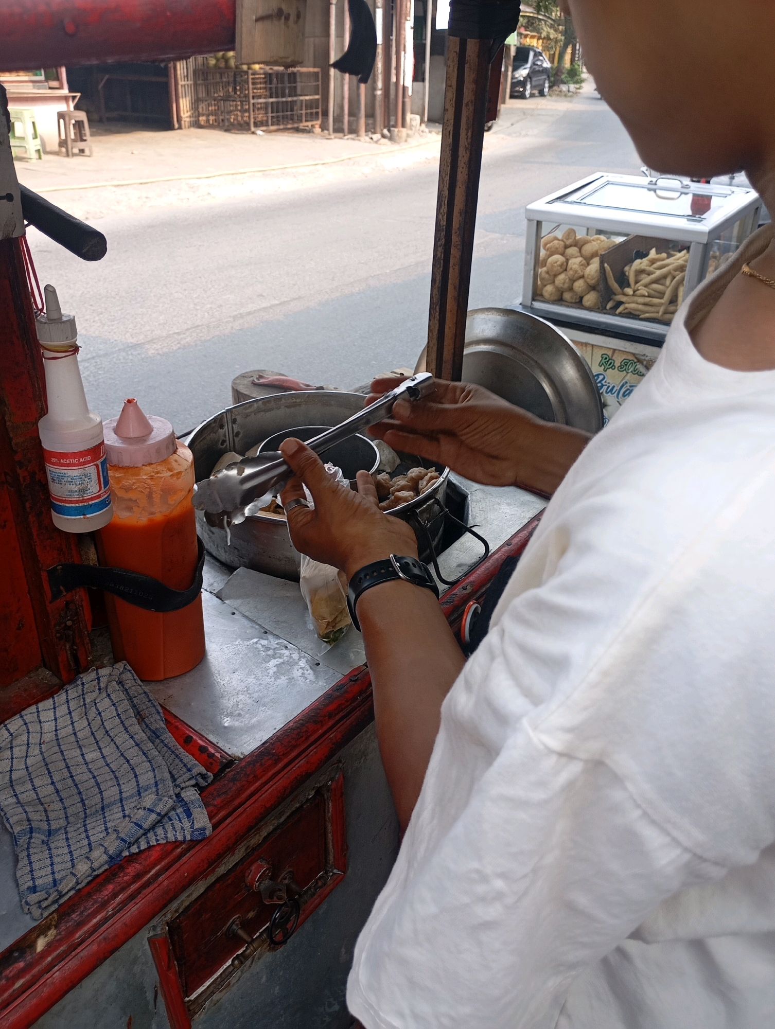 This is a medium shot, street-level photograph taken from the perspective of a vendor of a food cart in Babelan, Indonesia. A person wearing a white t-shirt is seen from the back, with their arms reaching towards a stainless steel pot containing what appears to be meatballs or dumplings. The vendor is holding a pair of tongs, actively serving the food. To the left of the pot are two plastic squeeze bottles, one with a white cap and orange liquid, labeled as "25% Acetic Acid," and another larger orange bottle with a pink cap, containing a reddish sauce. A blue and white checkered cloth is folded on the counter below the bottles. Behind the food cart, a street is visible with some buildings and a parked car in the distance. The cart itself is constructed with red painted wood and metal elements, showing signs of wear and tear. A glass display case with two compartments sits on the cart, one filled with round, light-colored items (likely meatballs) and the other with elongated fried snacks. A sign on the cart displays "Rp. 500..." indicating prices. The lighting suggests it is daytime.