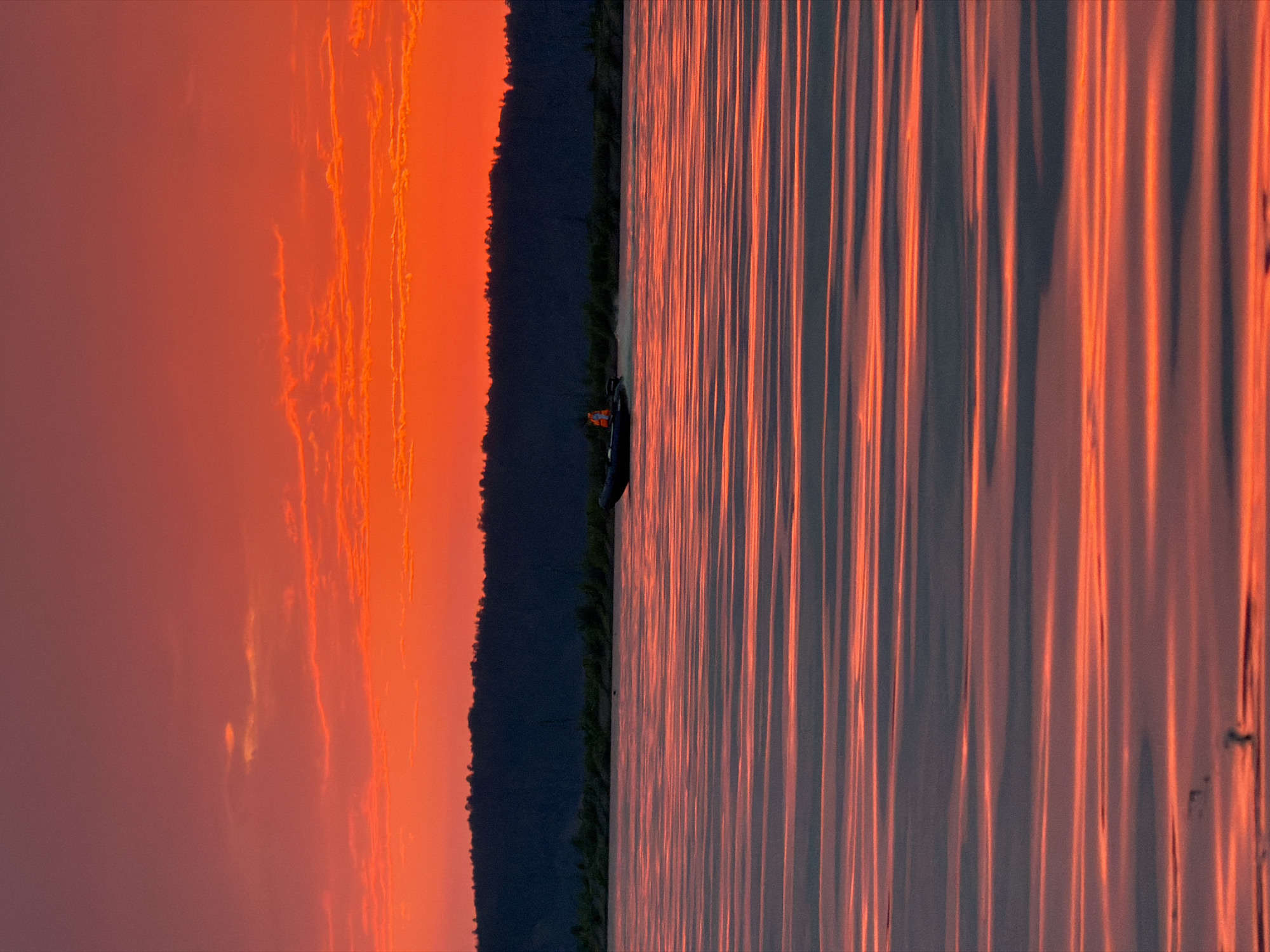 The image captures a vibrant sunset over the Kama River in Chaikovsky, Russia. The sky is ablaze with intense oranges and reds, with wispy clouds scattered across the horizon. A dark, silhouetted treeline forms the backdrop for the fiery sky. In the foreground, the water's surface is a canvas of rippling reflections, creating horizontal bands of deep red and gray. A small, dark boat with a person wearing an orange life vest is moving across the river, leaving a subtle wake. The overall scene is dramatic and tranquil, showcasing the natural beauty of the location at dusk.