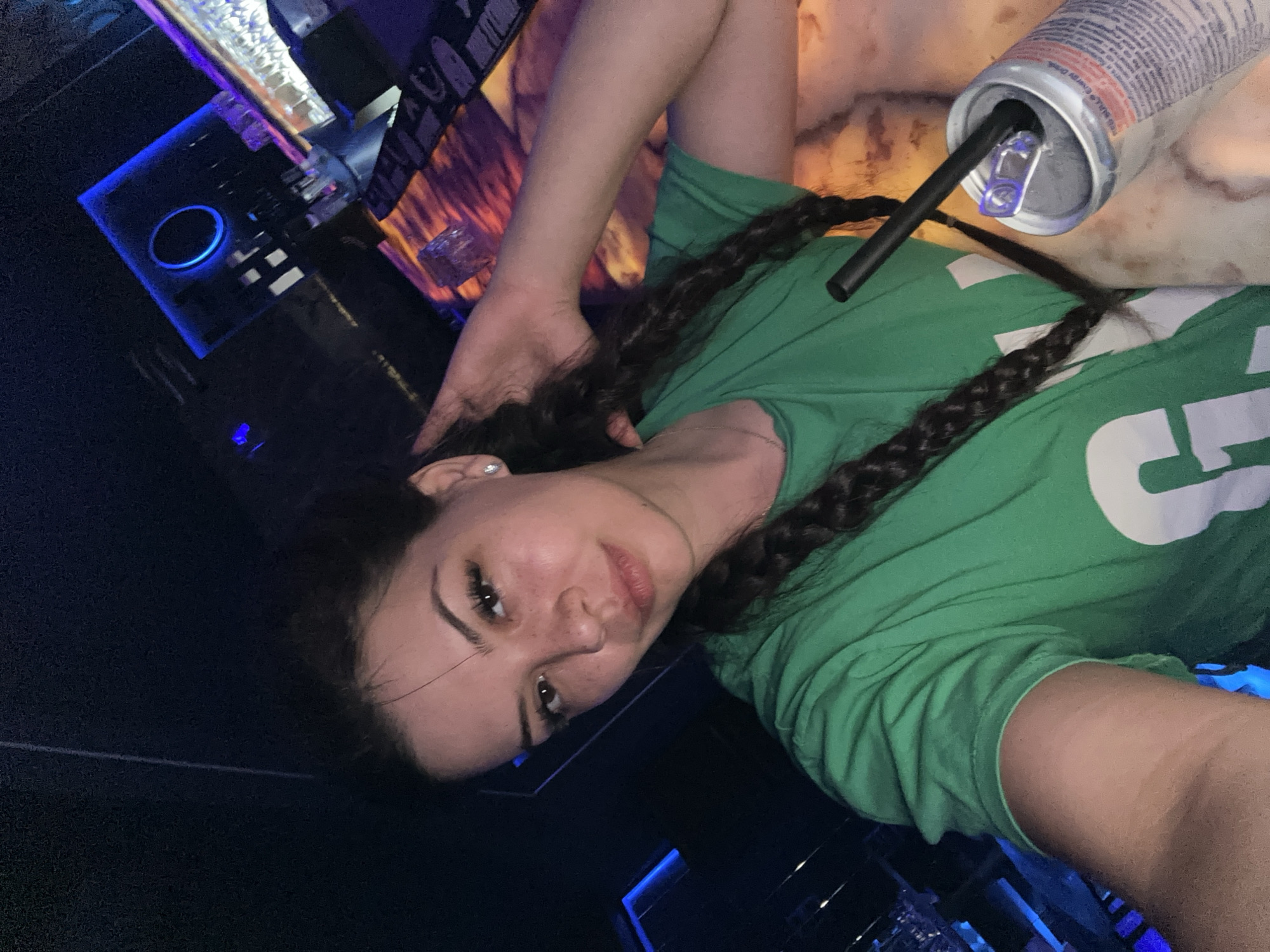 A young woman with dark hair in two braids and freckles on her face is taking a selfie in what appears to be a nightclub. She is wearing a green t-shirt and holding a can of Red Bull with a straw in it. The background shows a dimly lit bar area with blue and purple lighting. The woman is positioned in the foreground, with the bar and a counter visible behind her. The overall atmosphere suggests a nightlife setting. There is no indication of the specific location within the city of Sliven, Bulgaria.