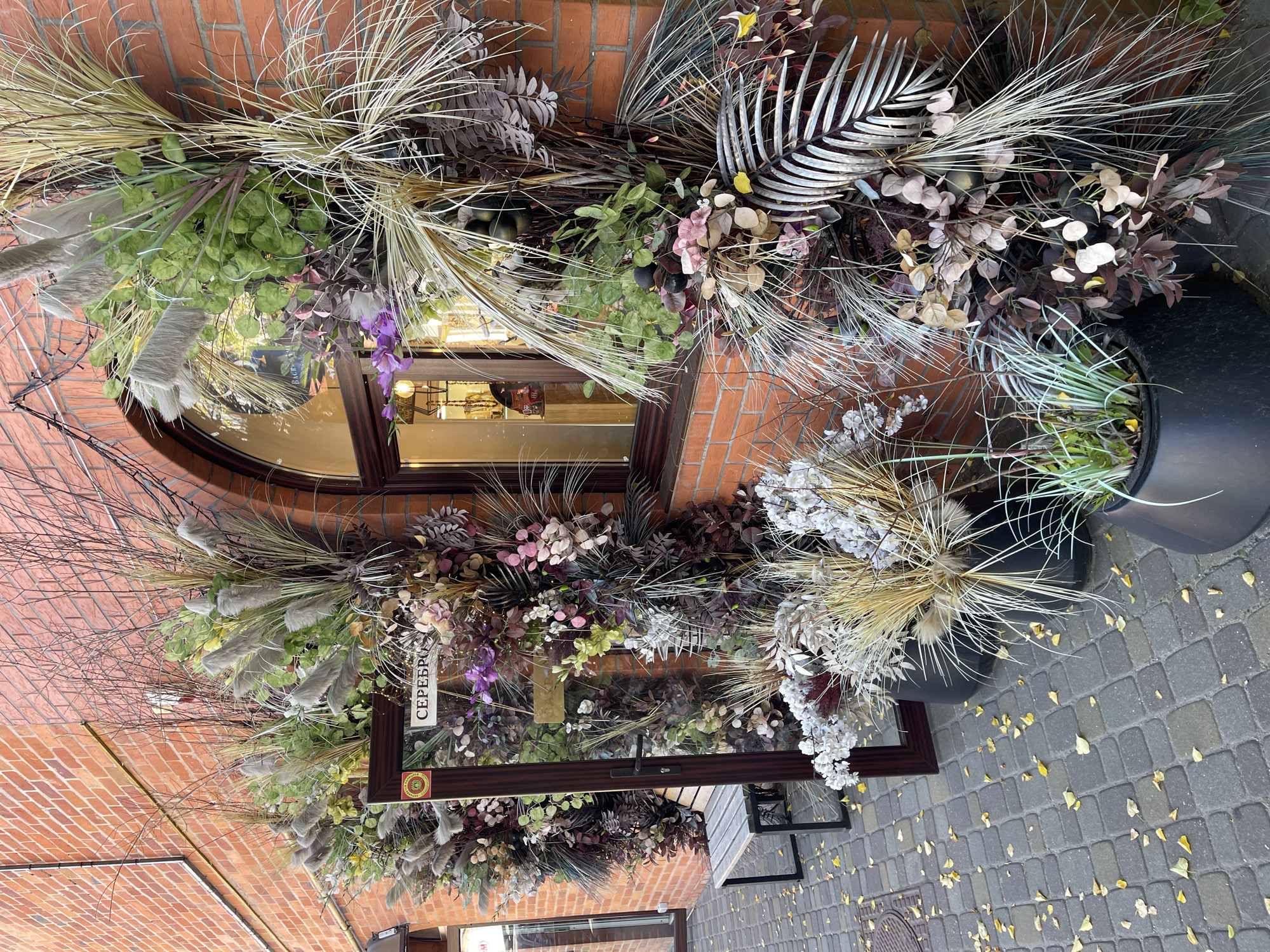 This media file captures an outdoor scene in front of a brick building. The foreground is dominated by an elaborate floral arrangement, featuring a mix of dried grasses, artificial foliage in various shades of green, purple, and beige, and delicate white flowers. The arrangement is quite dense and appears to be part of a shop's display, as a large window with a curved top is visible behind it. Reflections in the window suggest it might be daytime, with an overcast sky.
The floral arrangement itself is rich in texture and color, with tall, feathery plumes of grass interspersed with smaller, leaf-like elements and some darker, almost plum-colored leaves. A wooden frame, possibly part of a sign or display, is placed within the arrangement, and a small white label with text is attached to it.
The ground in front of the building is paved with gray interlocking stones, and scattered yellow leaves suggest an autumn setting. To the right of the frame, large black planters are visible, holding more of the dried grasses and foliage, further emphasizing the decorative nature of the display. The overall impression is one of a thoughtfully designed and seasonally appropriate storefront decoration.
The visible text on the label reads "СЕРЕБРЯН" which is likely part of a longer word in Cyrillic script. A circular emblem with yellow and red colors is also visible on the wooden frame. The setting appears to be a public walkway or sidewalk.