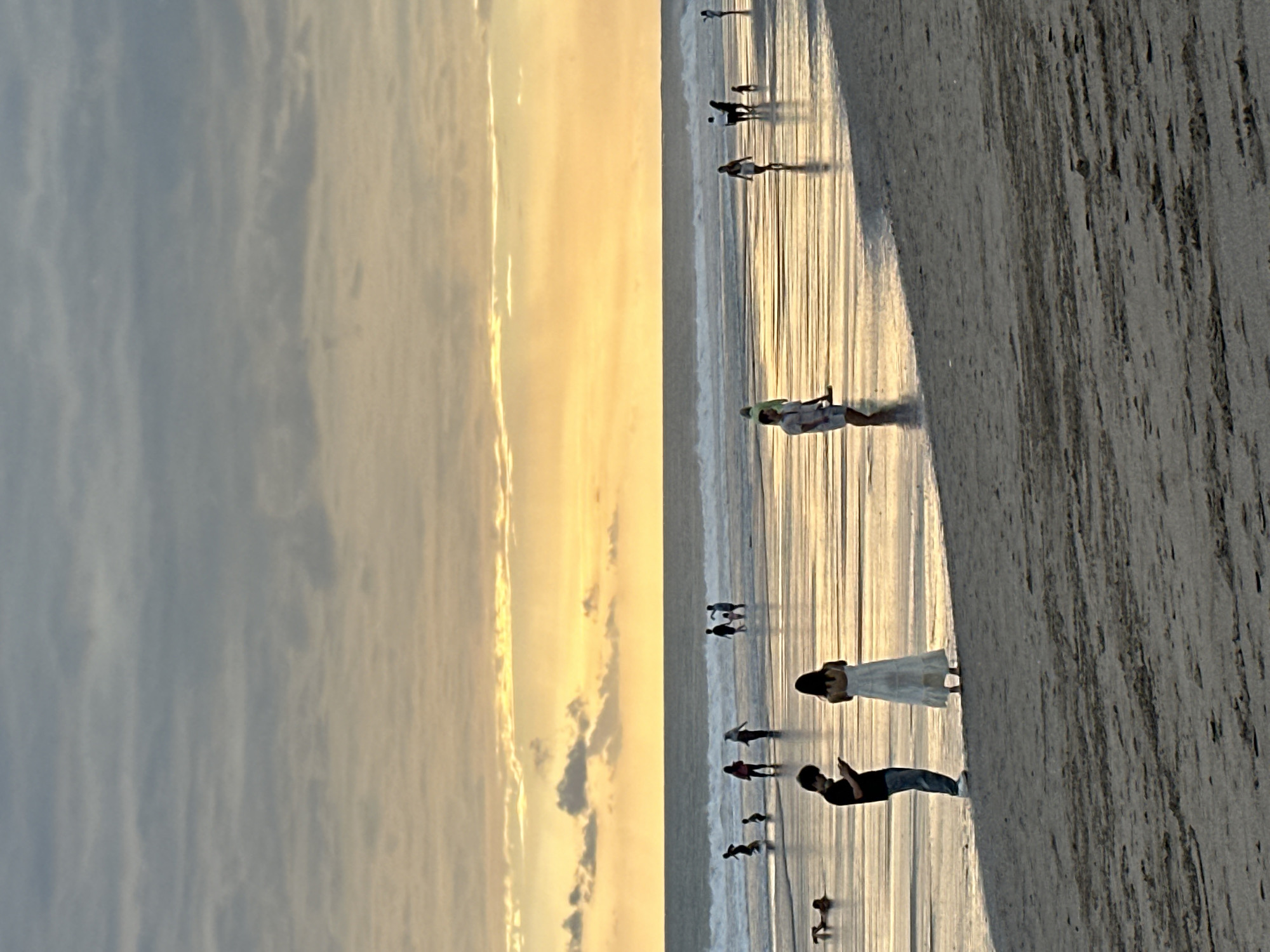 At sunset on a beach in North Kuta, Indonesia, several people are scattered across the wet sand and shallow water. A man in a black shirt and jeans stands looking at his phone, while a woman in a long white dress stands nearby. Further out in the water, a group of people are walking, some with children, and another individual is lying down near the shore. The sky is filled with warm orange and yellow hues from the setting sun, with scattered clouds. The wet sand reflects the colors of the sky, creating a shimmering effect. Waves gently break on the shore in the background.
