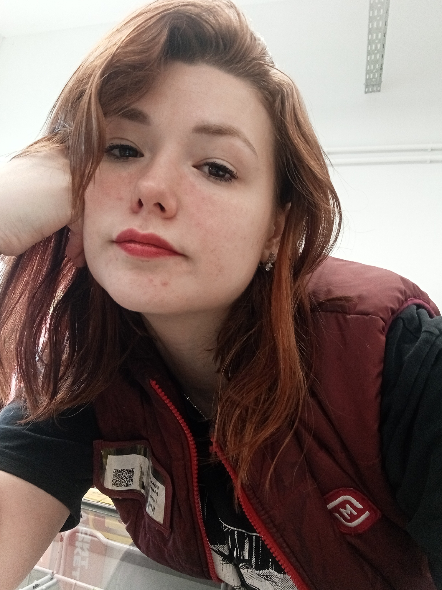 A medium close-up shot depicts a young woman with reddish-brown hair, fair skin, and light freckles. She is wearing a maroon puffer vest over a black long-sleeved shirt. The vest has a patch on the right side and a QR code in a plastic sleeve on the left. She wears small stud earrings. Her makeup includes reddish lipstick. She rests her head on her left hand. The background includes a white ceiling with a metal track lighting system and a white wall. The image was taken at an elevated angle. There are no readily apparent visual cues in the photograph to definitively place the subject in Chaikovsky, Russia. The QR code on her vest might reveal more information, but it cannot be determined with the given information.