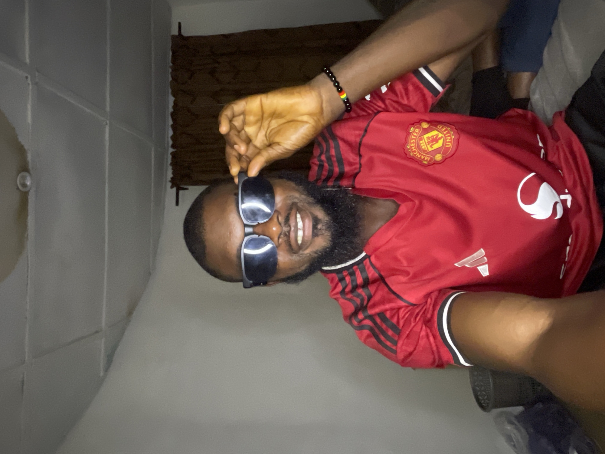 In a dimly lit indoor setting, a Black male is captured in a selfie. He is wearing a red Manchester United jersey with black Adidas stripes on the sleeves. The jersey features the Manchester United crest on the left chest and a white Adidas logo on the right. He has a beard and is wearing dark sunglasses, which he is adjusting with his right hand. On his left wrist, he wears a black beaded bracelet with colorful bands, including red, yellow, and green, possibly representing the Pan-African flag. The background shows a plain wall, a dark brown curtain, and part of a white ceiling with visible paneling and a light fixture. To the left, a dark-colored trash bin and some fabric items are visible. The overall lighting suggests the photo was taken in a residential area within Jos, Nigeria.