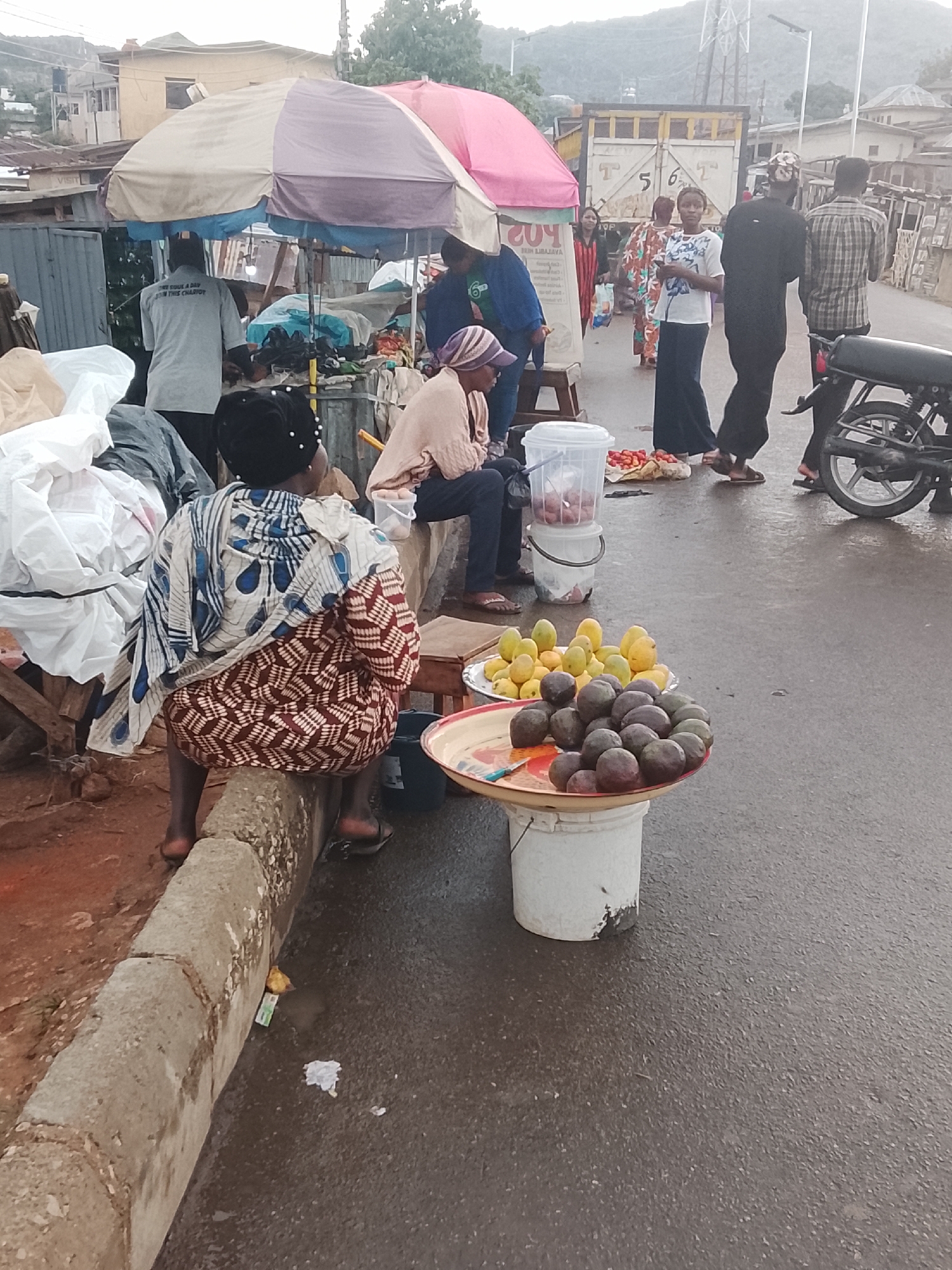 The image depicts a street scene in Gwong, Nigeria. A woman in a patterned dress sits on the edge of a concrete curb, facing away from the camera. Next to her is a tray of dark purple fruits resting on a white bucket. Another tray of yellow fruits is visible behind. An assortment of people and goods are under a multi-colored umbrella, indicating a makeshift market setup. A man in a grey shirt stands under the umbrella, his back to the camera. People are walking on the street, and a motorcycle is parked on the right side of the frame. A truck is visible in the background. The overall impression is of a bustling, informal market area on a wet-looking street.