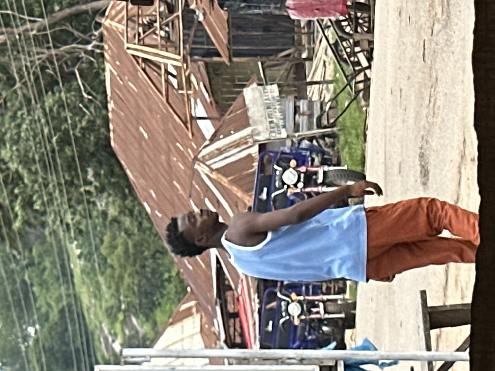 In Mararaba, Nigeria, a young Black man wearing a blue tank top and orange pants stands in profile, looking to his right. He is positioned in the foreground of the image. Behind him, several buildings with weathered, corrugated metal roofs are visible. Two blue and red three-wheeled vehicles are parked near the buildings. In the background, lush green trees and foliage are present, with power lines stretching across the scene. The ground is a light-colored, dusty surface, possibly a road or pathway. A wooden bench is partially visible in the lower left of the frame. A metal handcart with large spoked wheels is on the right side of the image, and a piece of pink fabric is draped over it. The overall impression is of a busy, perhaps somewhat rural or semi-urban, environment.