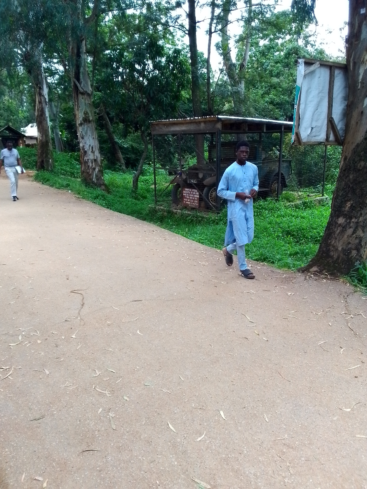 A man in a light blue tunic and matching pants walks on a dirt path that curves to the left. He is wearing dark sandals. Behind him, a second man in a gray t-shirt and white pants walks towards the left. In the background, there are trees, lush green foliage, and a covered exhibit that appears to be displaying an old military vehicle. The exhibit is surrounded by a fence. The environment appears to be a park or outdoor museum.