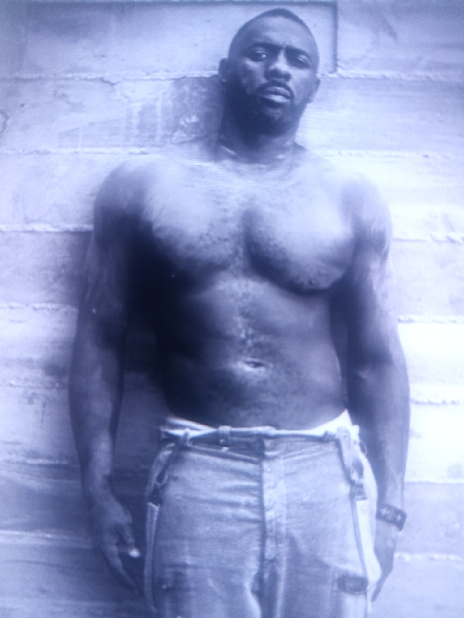 A shirtless man with dark skin, a shaved head, and a goatee stands facing forward, leaning against a textured wall. He is wearing light-colored pants with suspenders. The man's chest and arms are muscular. The background wall appears to be made of horizontally stacked bricks or stones. The image has a blueish tint and appears to be a photograph.