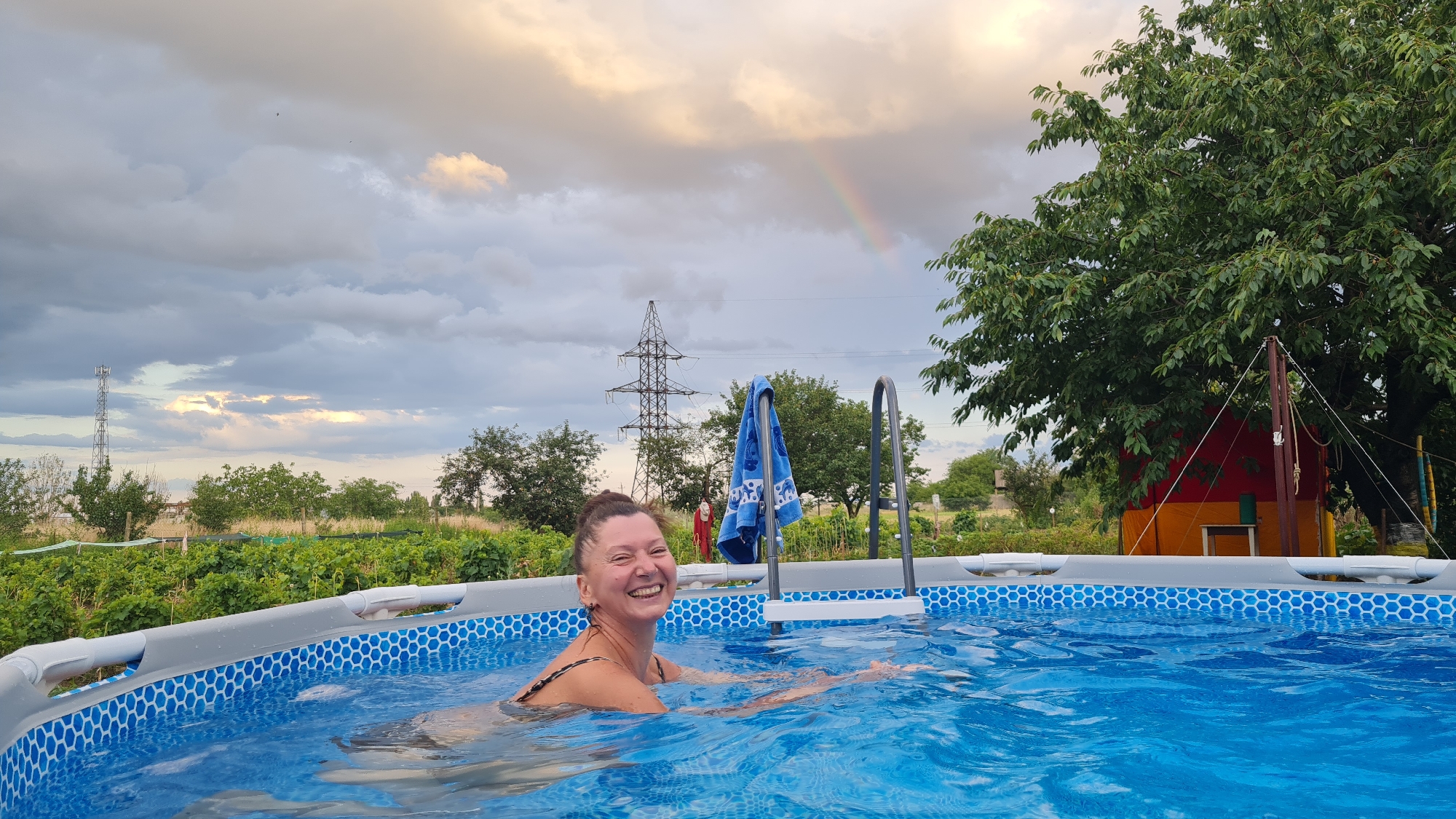 In Salhany, Ukraine, a woman is pictured in an inflatable swimming pool. She is smiling and looking at the camera. The pool has a blue hexagonal pattern around its edge and is filled with clear blue water. A blue towel hangs from the ladder of the pool. In the background, there are trees, a vineyard, and a power line tower. A faint rainbow is visible in the cloudy sky.