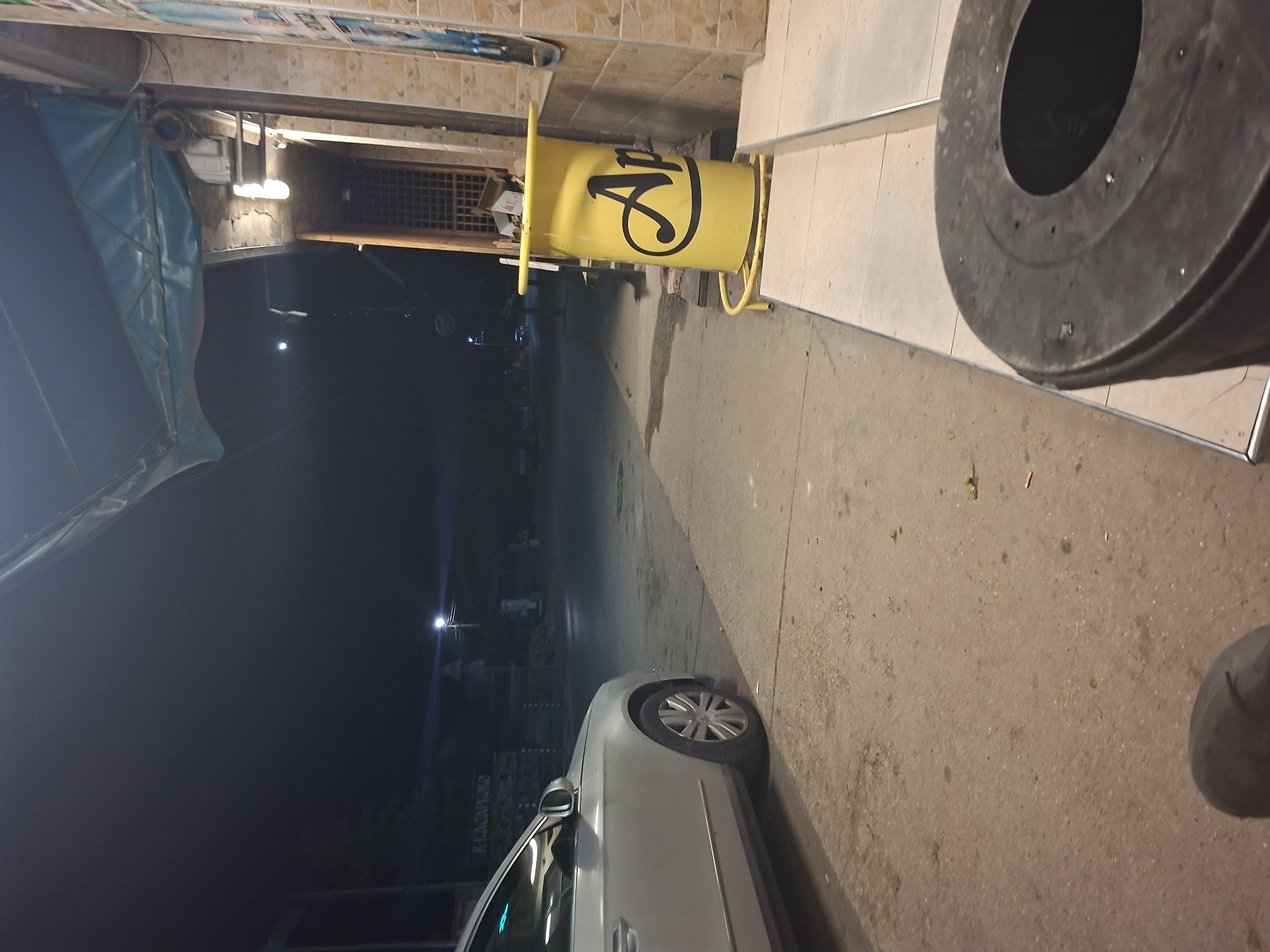 This nighttime image depicts a street scene in Basarbovo, Bulgaria. In the immediate foreground, a dark, cylindrical outdoor ash bin or trash receptacle is visible on the right, with a small portion of a dark-colored shoe or boot on the lower left. The ground consists of a concrete sidewalk with visible dirt and scattered small debris, including what appears to be cigarette butts and green organic matter.
To the right, a tiled building facade with light brown/beige square tiles is present, featuring a light blue/teal awning extending over the sidewalk. Mounted beneath the awning are bright linear light fixtures. Adjacent to the building and on the sidewalk is a yellow, cylindrical stand or dispenser with black lettering, partially obscured, which reads "Ap" (likely "Apost").
On the left side of the concrete sidewalk, a light-colored (possibly silver or white) sedan is parked, with its front section extending out of frame to the left. Its front passenger wheel and side profile are clearly visible. Beyond the car and across the street, the paved road is dark and mostly empty, leading into the distance. Faintly visible on the left side of the distant street is a sign or structure with the Cyrillic text "БРАКАПОГОЛО". Several bright streetlights illuminate portions of the distant road, creating noticeable light flares. The sky above is dark, indicating a clear night. No individuals are explicitly visible beyond the implied presence of the photographer.