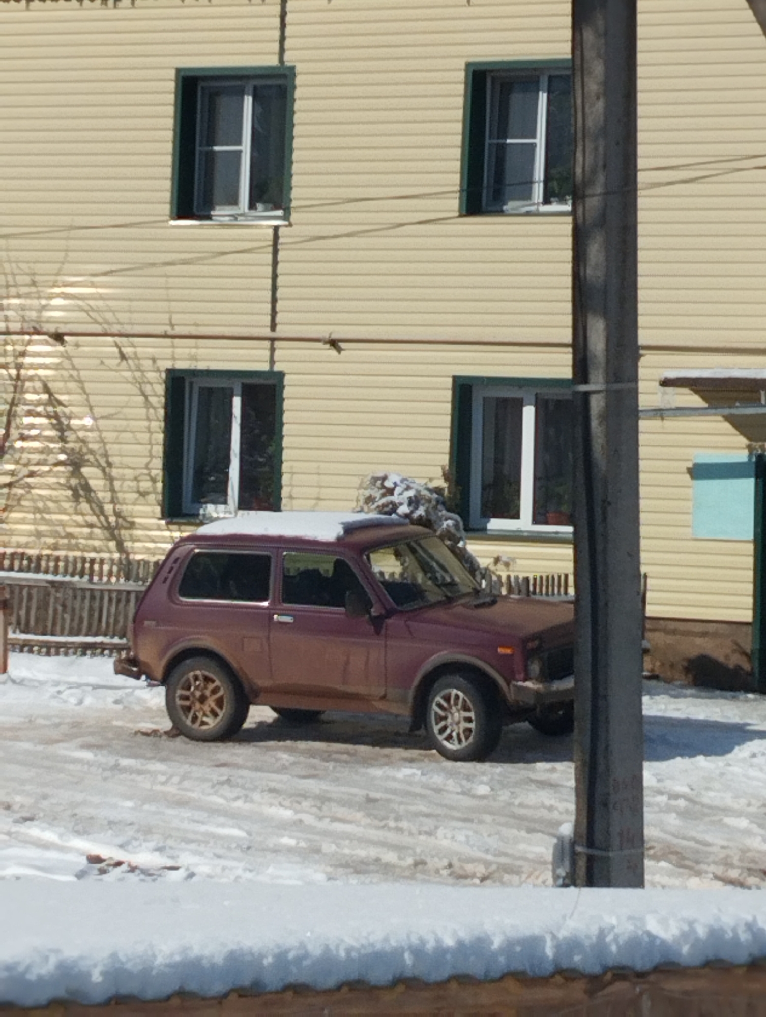 The image depicts a muddy red Lada Niva parked in a snowy residential area of Krasnogorskoye, Russia. The car has snow on its roof. Behind the car is a light yellow two-story house with four windows. A wooden fence partially obscures the view of the house. A utility pole stands in the foreground to the right. The ground is covered in snow and patches of mud.