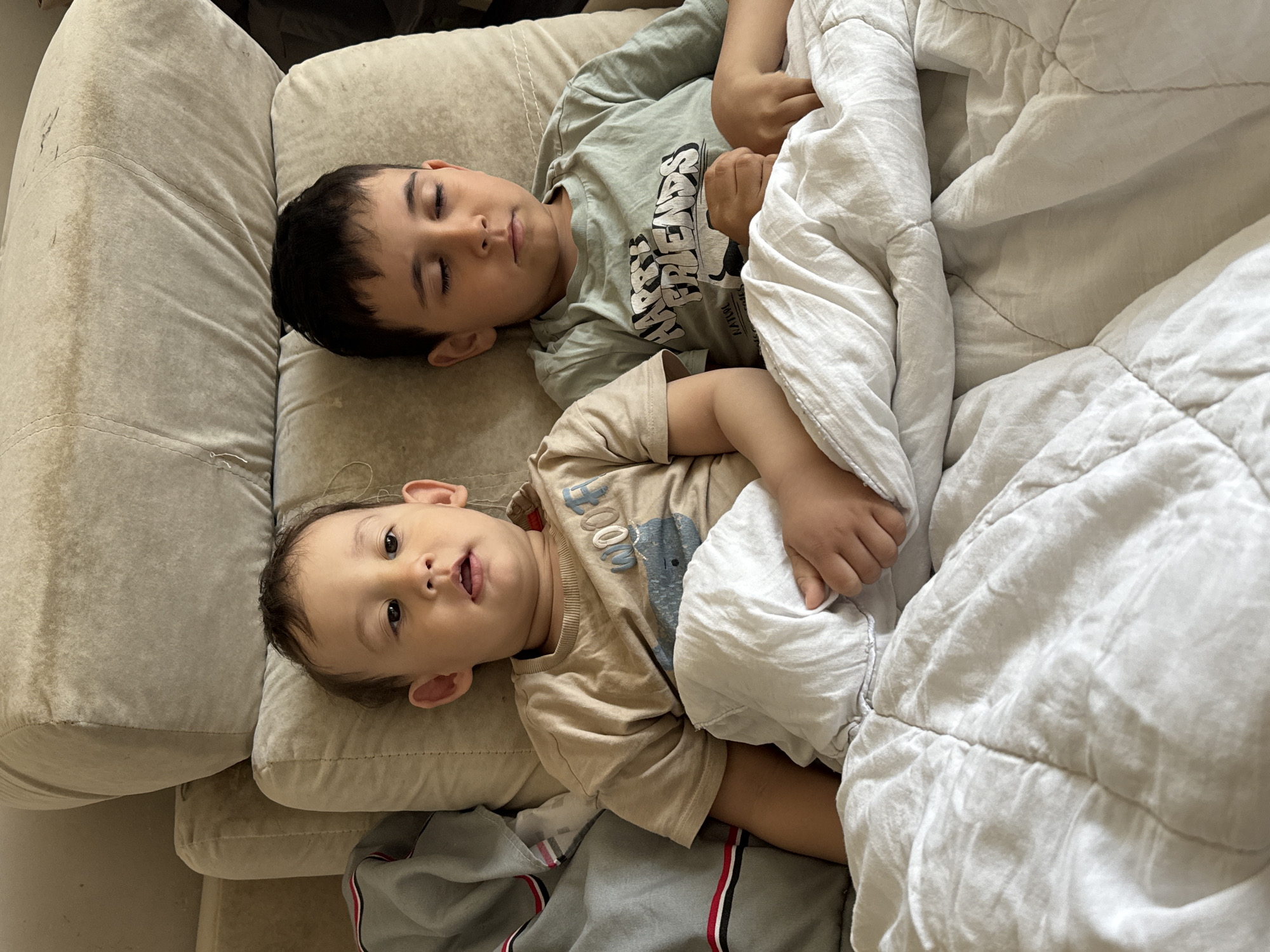 Two children are lying on a beige sofa under a white blanket. One child, likely older, is asleep with dark hair and wearing a green t-shirt. The second child, possibly a toddler, is awake and looking at the camera, wearing a tan t-shirt. Part of gray pants with red and white stripes are seen near the toddler. The location is likely indoors, but there is no confirmation that it is in the city of Chaikovsky, Russia.