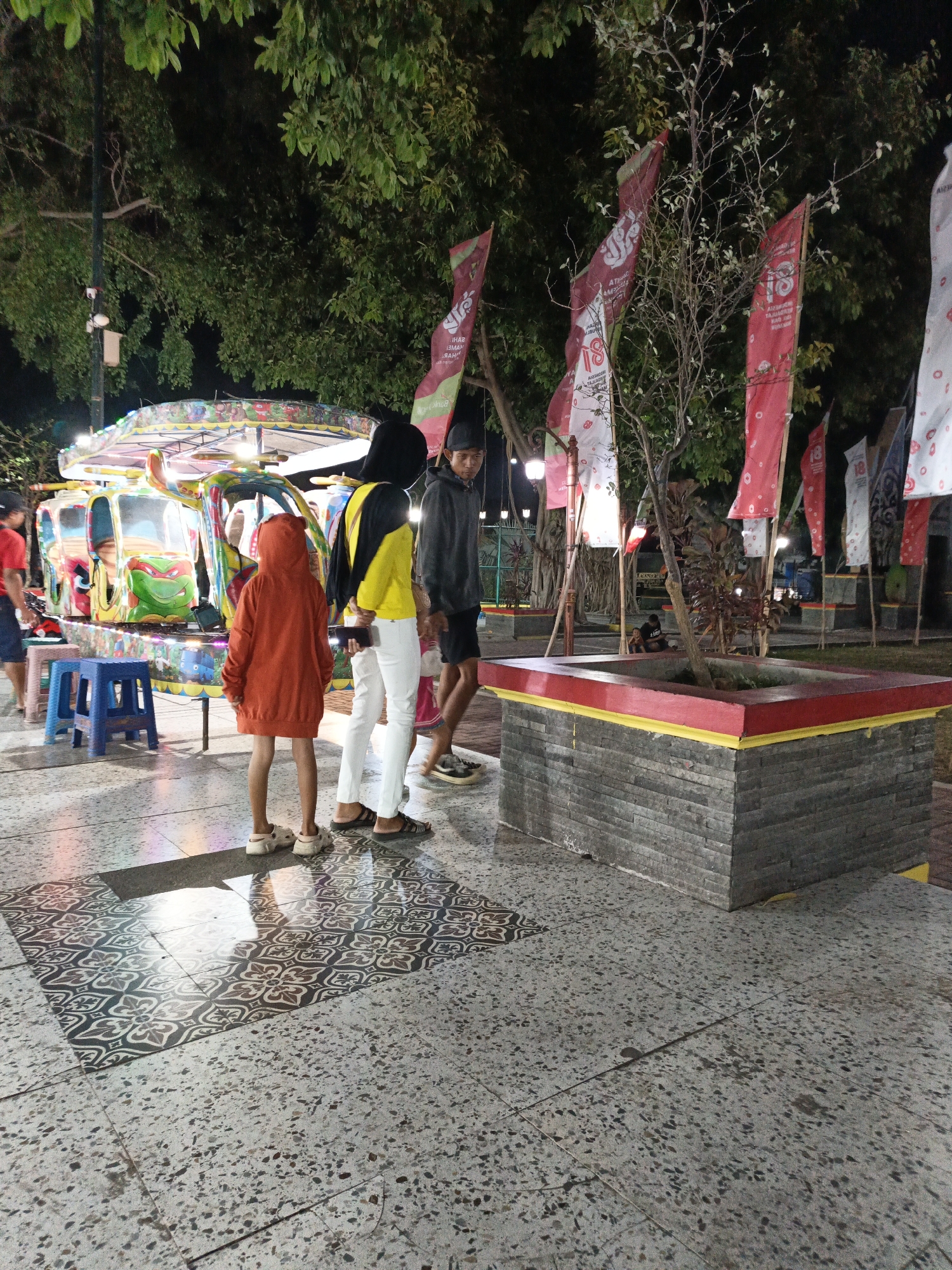 The image depicts an outdoor scene at night, possibly in a park or recreational area in Klaten, Indonesia, as indicated by the presence of decorative flags and a colorful amusement ride. In the foreground, a person wearing an orange hooded jacket and white sneakers stands facing away from the camera, with their hands at their sides. To their right, another individual in a bright yellow shirt, black hood, white pants, and sandals is also observed from behind. A third person, seen from the side, wears a black cap, a dark gray hoodie, and dark shorts, with a concerned expression.
Dominating the mid-ground is a vibrant, multi-colored amusement ride, possibly a carousel or a similar attraction, illuminated by lights. In the background, several tall, slender flags are displayed; some are red with white markings and the number "18," while others are white with red circular patterns. A potted plant with bare branches is situated in a stone planter box with red and yellow trim. The ground is paved with speckled concrete tiles and features a section of patterned tiles in the center foreground. Trees with dense green foliage are visible, partially obscuring the night sky. The overall ambiance suggests a lively nighttime gathering or event.