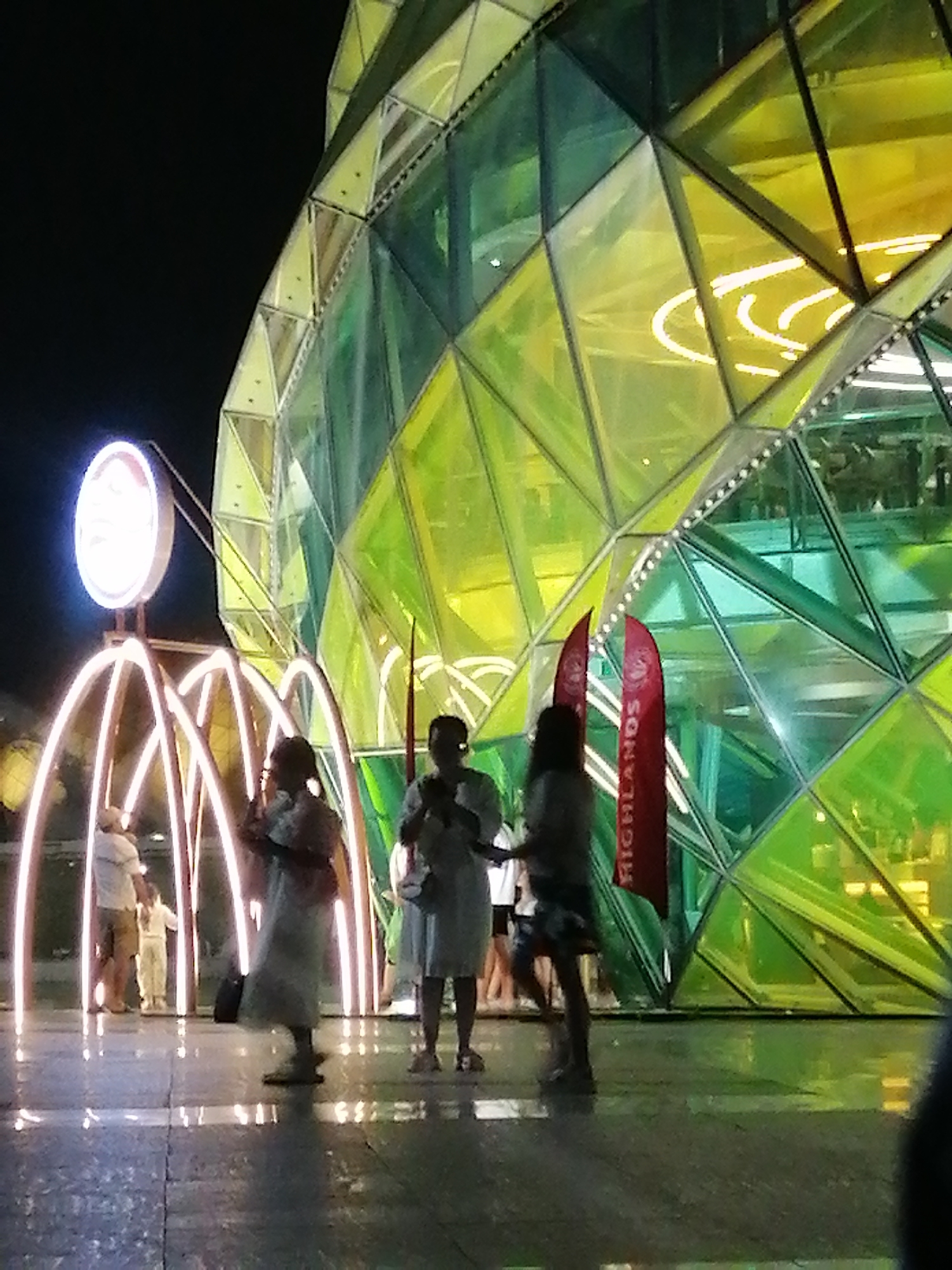 The image depicts a nighttime scene in Da Lat, Vietnam, focused on a modern, geometrically faceted building with translucent green and yellow panels. In the foreground, several people are visible on a wet, reflective tiled surface. To the left, a child stands near a man in a white shirt and shorts, both illuminated by arching, glowing light structures. Further right, three individuals are standing, two of whom appear to be women. One woman in the center wears a light-colored dress and holds a dark bag. Another woman on the far right is partially obscured and carries a bag. Two red flags with white text, one reading "HIGHLANDS", are positioned behind the central figures. The overall atmosphere suggests an outdoor public space, possibly a plaza or promenade, adjacent to a distinctive architectural structure.