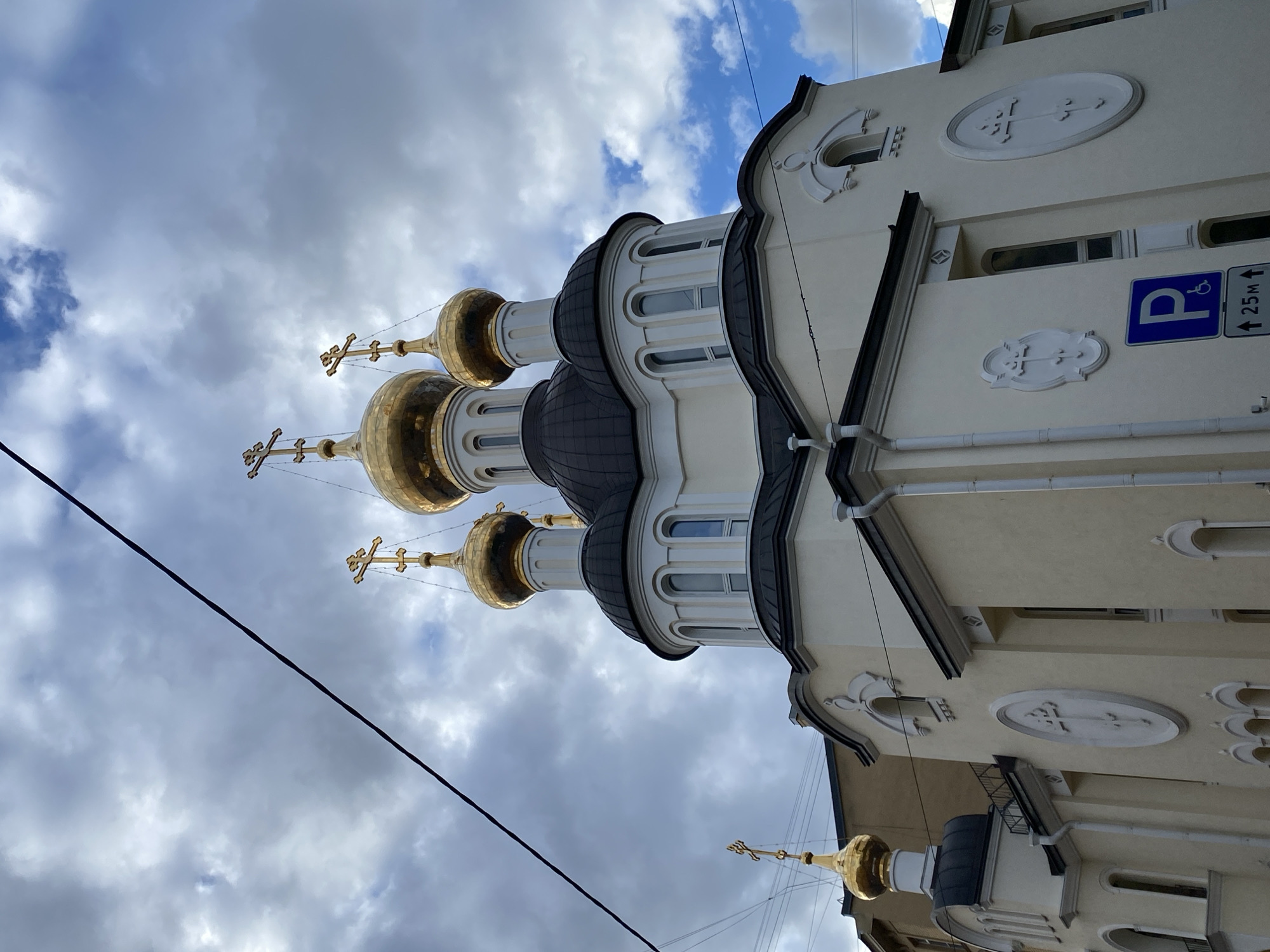 The image shows a low-angle view of a Russian Orthodox church in Saint Petersburg, Russia. The church is characterized by its multiple golden domes topped with Orthodox crosses. The building's facade is light-colored, with arched windows, decorative moldings, and religious symbols. A parking sign for disabled individuals is visible on the side of the building. Overhead, a cloudy sky fills the background, and a power line runs diagonally across the frame.