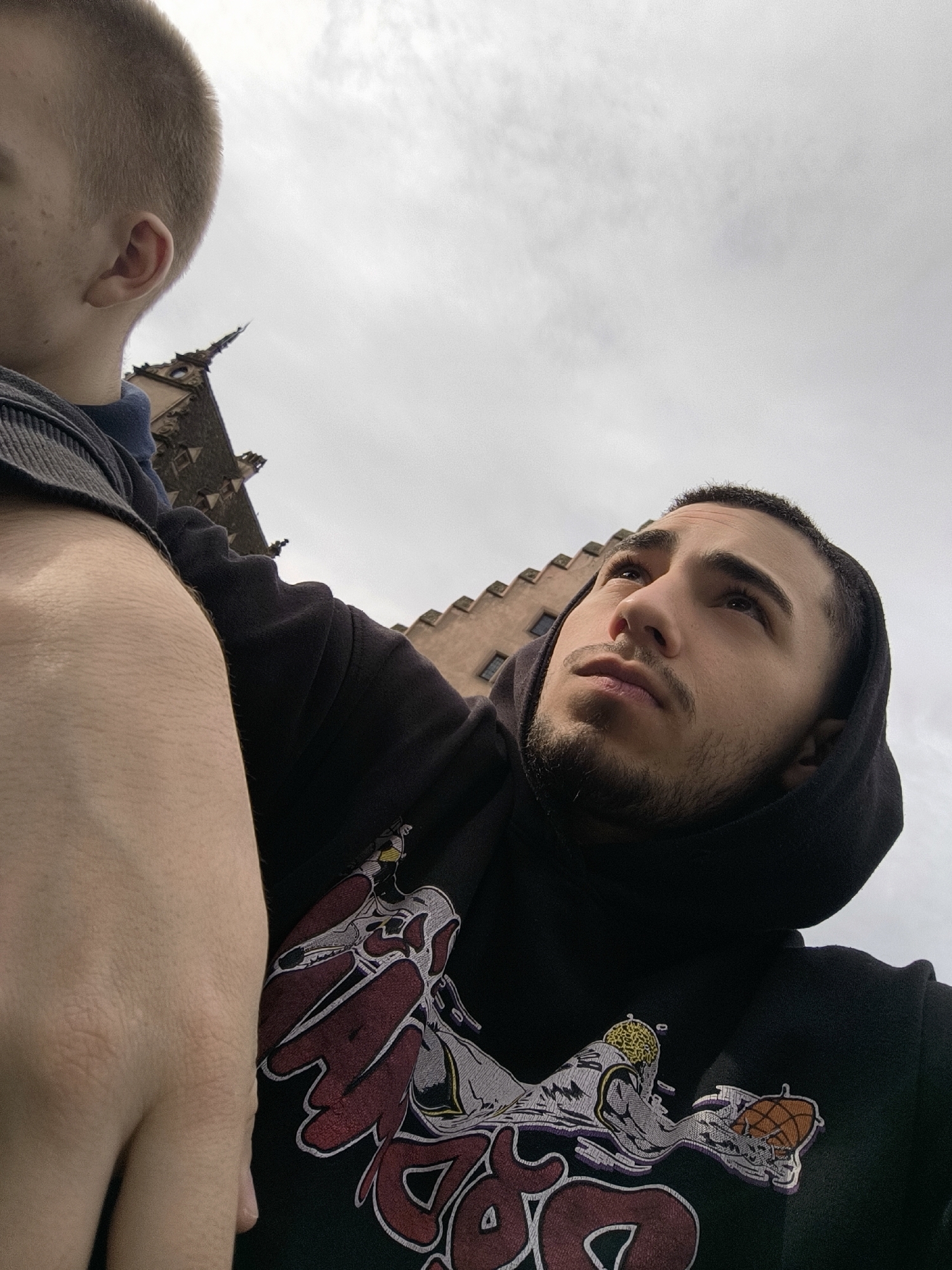 The image is a low-angle shot of a man with short dark hair and a beard, wearing a black hoodie with a large graphic design featuring a basketball player and stylized text. To his left is another person, cropped at the head and shoulder, with short blond hair. Behind them is a building with a pointed roof and visible windows, partially obscured by clouds. This building is likely part of the Strasbourg Cathedral, placing the location in Strasbourg, France. The camera angle suggests the people are looking up at something. The lighting is overcast, indicating a cloudy day.