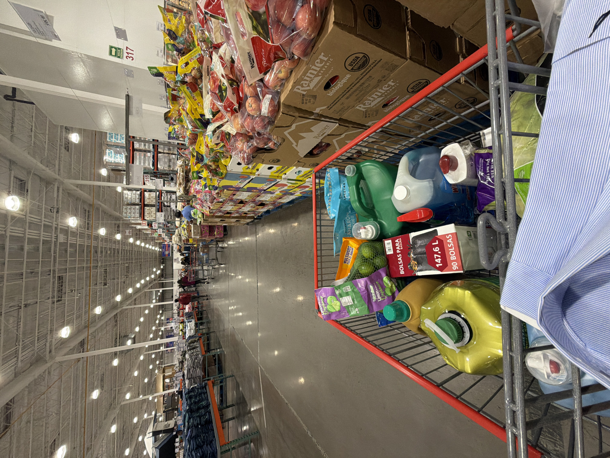 The image shows a shopping cart in a Costco-style store in an unidentified location. The cart is filled with various items, including boxes of Rainier Gala apples, bags of "chomis" (likely a snack), trash bags ("BOLSAS PARA BASURA"), cleaning products, and other grocery items. The aisle is lined with similar products on shelves, and shoppers can be seen in the distance. Signs in the background include the number "317" and "315" and the word "EVACUACION". The lighting is bright and the environment appears to be a large retail warehouse. There is also a glimpse of a blue-striped shirt on the right side of the frame, presumably worn by the person pushing the shopping cart.