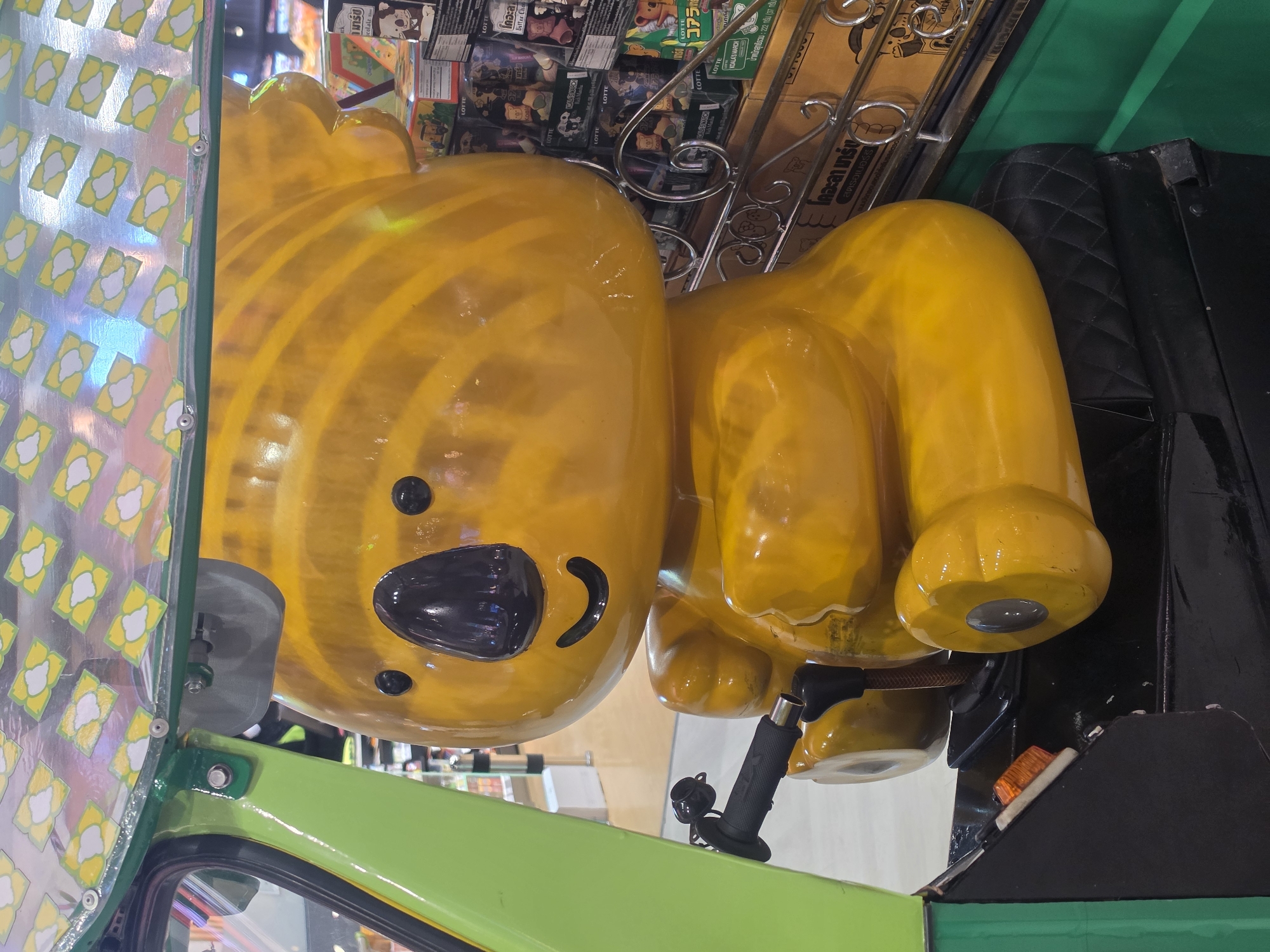 An interior shot, taken in Bangkok, Thailand, features a large, glossy yellow cartoon character, resembling a smiling koala or bear, dominating the frame. The character has black eyes, a black nose, and a curved black line forming a happy mouth, with concentric ridges on its body. It appears to be a decorative element or mascot, possibly made of plastic or fiberglass.
The character is situated within what seems to be the cabin of a vehicle, likely a modified tuk-tuk. Green painted surfaces are visible on the lower left and bottom, forming part of the vehicle's structure. On the right, a black, quilted-patterned material suggests seating or interior paneling. A black handlebar with a rubber grip is seen near the character's right "arm," and an orange indicator light is visible on the black structure.
To the left, a translucent panel decorated with repeated yellow and white stylized floral patterns reflects light from an unseen source. In the background, behind the yellow character, shelves are stacked with numerous small boxes of "Lotte Koala's March" cookies, many featuring cartoon koala illustrations. Visible text on the packaging includes "LOTTE," "KOALA'S MARCH," "コア" (Koala in Japanese Katakana), "มาร์ช" (March in Thai), "สตรอเบอร์รี" (Strawberry in Thai), and "Bitter Chocolate." A metallic, scrolled railing is also present among the boxes. The scene is static, showcasing the character and its immediate surroundings.