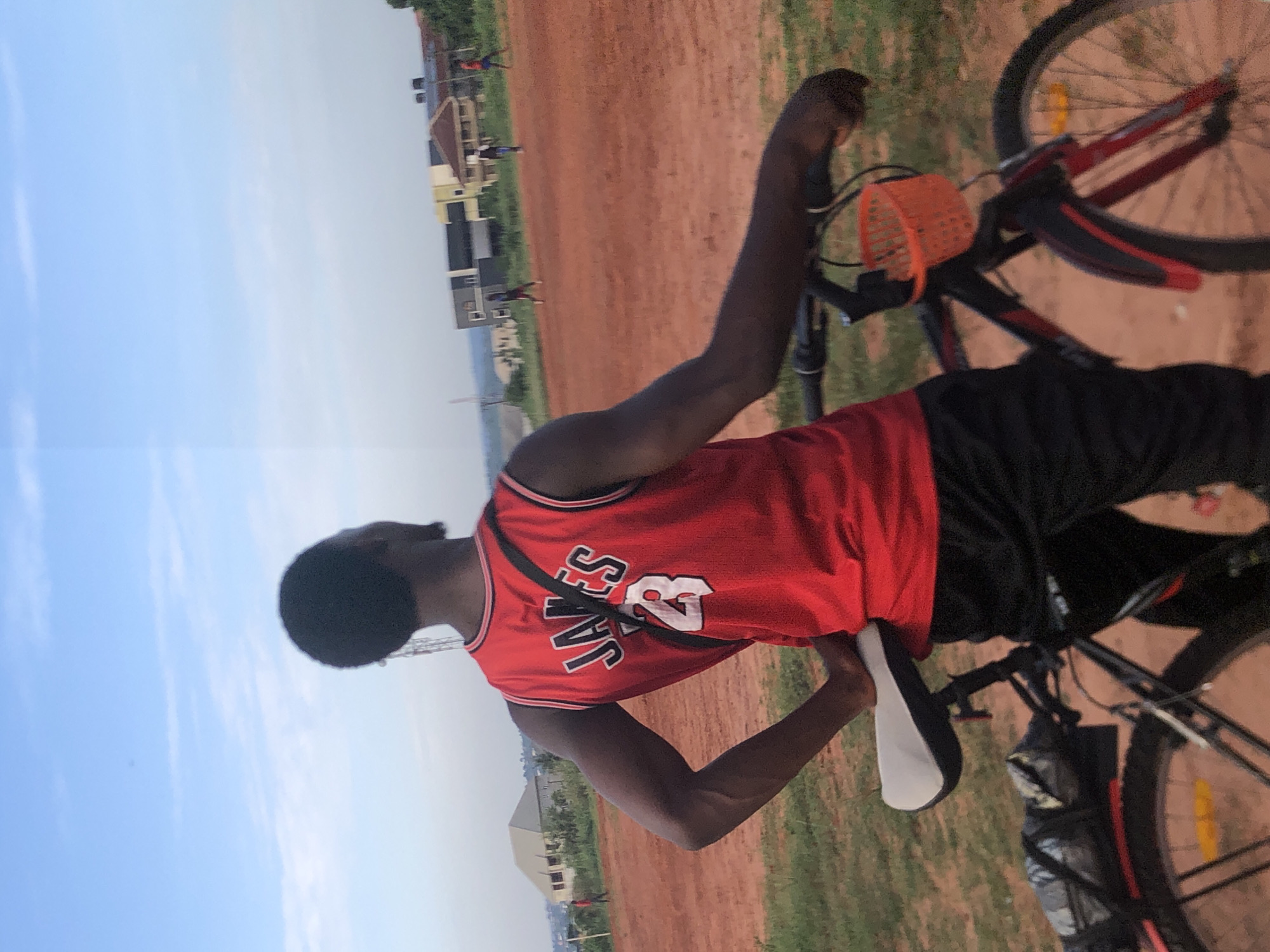 This outdoor photograph, taken in Ukwu-Abwa, Nigeria, features a young individual and their bicycle in an open field under a clear sky. The image is captured from behind the person, who is facing away from the camera and slightly to the left.
The main subject is a person with dark skin and short dark hair, wearing a red sleeveless jersey that has "JAMES" and the number "23" printed in white with a black outline on the back. They are also dressed in black shorts or long pants, and a black strap, possibly from a shoulder bag, is visible across their back.
Beside the person is a bicycle with a black frame accented with red details on its fenders. An orange, semi-transparent basket is attached to the handlebars. The bicycle also features a white and black saddle and a partially visible wheel with a yellow spoke reflector.
The ground consists of reddish-brown soil intermingled with patches of green grass, characteristic of many open fields in the region. In the background, several light-colored buildings of different sizes can be seen, suggesting a residential or semi-urban environment. Distant, blurred figures of other people are visible, some appearing to be walking or engaging in activities.
The sky is a bright, clear light blue with a few faint white cloud streaks, indicating a daytime setting, likely late morning or afternoon. The scene conveys a casual, peaceful moment in a community area.