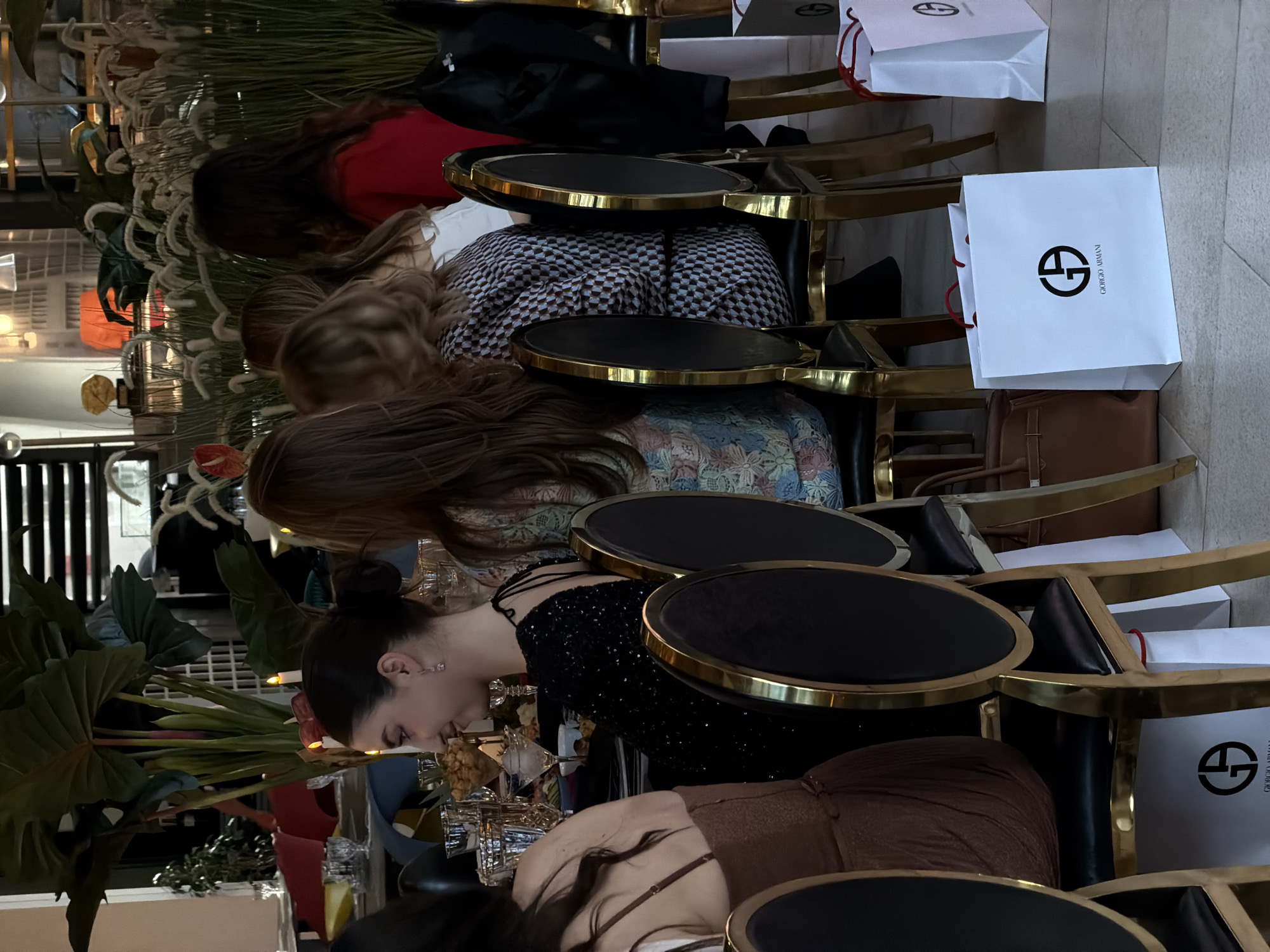 The image shows a group of people sitting at a table, likely in a restaurant or cafe in the city of Budva, Montenegro. The table is decorated with plants and glassware. The patrons are seated in chairs with gold frames and black seats. Some of the customers have shopping bags, particularly Giorgio Armani bags, placed by their chairs. They appear to be mid-meal or conversation, with a general focus on the table.