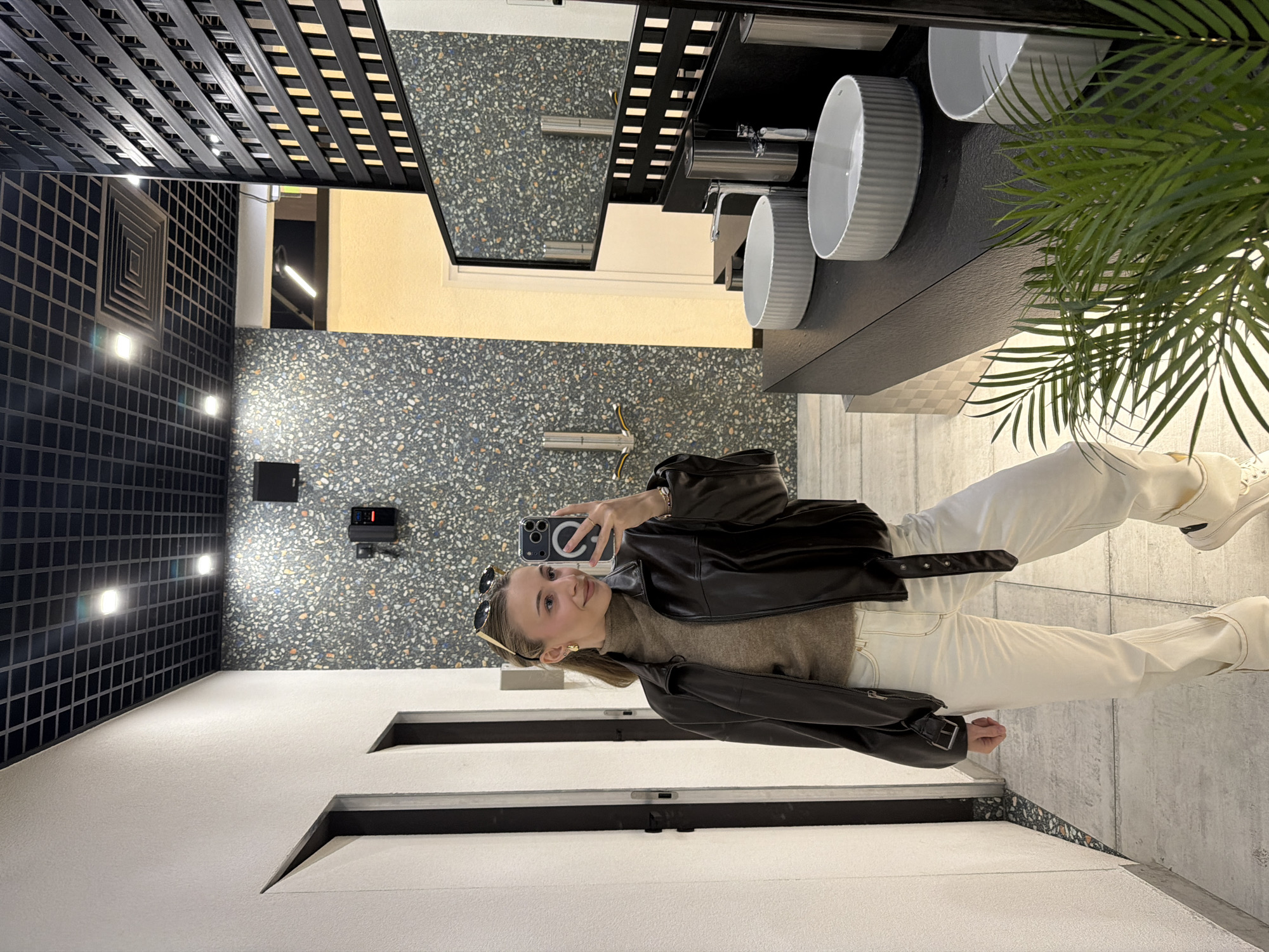 A young adult female is depicted taking a mirror selfie within a modern public restroom environment in Plovdiv, Bulgaria. She stands facing the camera, positioned slightly to the left of the frame's center.
**Person:** The female subject has light skin, light brown hair, and wears small gold hoop earrings. Sunglasses are perched atop her head. She is dressed in a brown turtleneck sweater, an oversized dark brown leather jacket, white wide-leg trousers, and white sneakers with black accents. Her left hand holds a black smartphone, which has a circular light attachment, aimed at the mirror. She exhibits a subtle smile.
**Environment:** The restroom features a dark gray terrazzo-patterned wall on the far side, flecked with white, black, orange, and blue elements. A black speaker and a control panel with red digital elements are mounted on this wall. To the left, a silver Dyson-style hand dryer is visible. The left wall is off-white and textured, featuring two dark-framed door openings. The ceiling is a black grid system with recessed square LED lights and a central square air vent. The floor is light gray with a subtle textured pattern.
**Objects:** A black minimalist vanity unit occupies the right side of the frame, supporting three white, ribbed, oval-shaped basin sinks. Each sink has a slender silver faucet, and silver cylindrical soap dispensers are positioned between them. A large rectangular mirror with a black frame is mounted above the vanity, reflecting the terrazzo wall and ceiling. A tall, vertical black lattice-style divider stands to the right of the subject. A green, palm-like plant is situated on the floor in the foreground, partially obscuring the vanity.