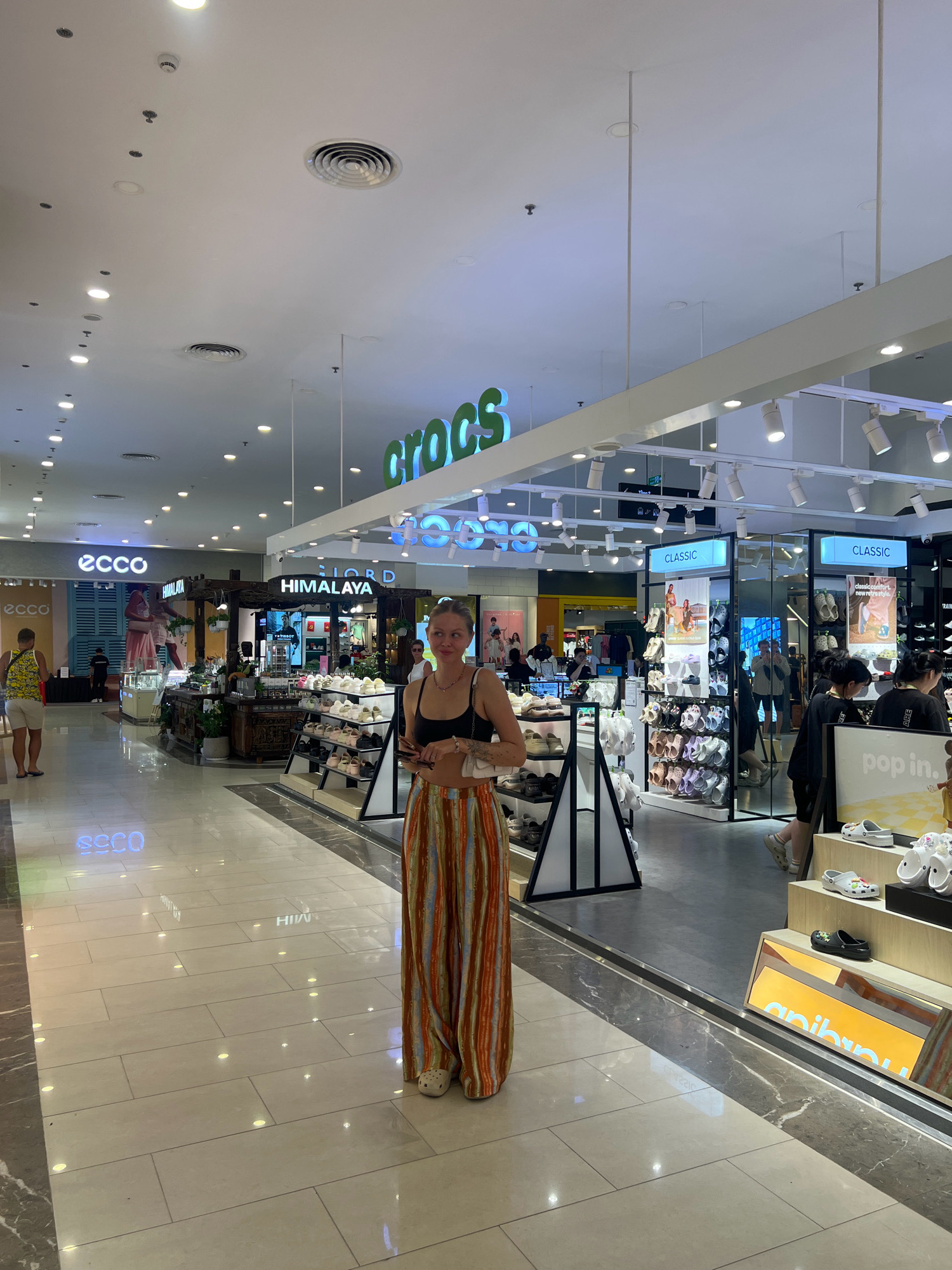 The image depicts the interior of a shopping mall in Nha Trang, Vietnam. A young woman with blonde hair, wearing a black crop top and brightly colored striped pants, stands in the foreground looking to the right. She is wearing white Crocs. Behind her, the mall hallway opens into a retail area. To the left, a store labeled "ECCO" is visible. Directly behind the woman is a store with a "HIMALAYA" sign and a display of various accessories. Further back and to the right, a "Crocs" store is prominently displayed with its logo in green neon letters. Multiple rows of Croc shoes are displayed on shelves within the store. Other shoppers can be seen browsing in the background. The mall features a polished tiled floor, overhead lighting, and a clean, modern aesthetic.