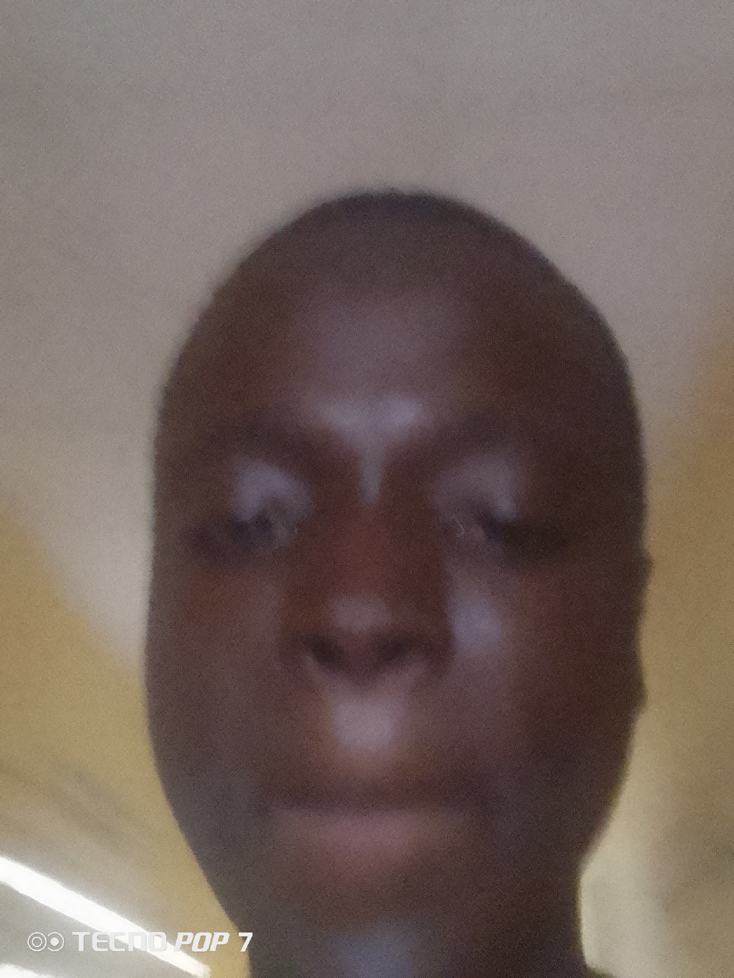 The image is a close-up, blurred photograph of a person's face, centered in the frame. The subject has dark skin and is looking directly at the camera. The background is a plain, light-colored wall. The bottom left corner of the image contains a watermark that reads "TECNO POP 7". The lighting appears to be artificial and casts some shadows on the face. The image's location is identified as Ipetumodu, Nigeria.
