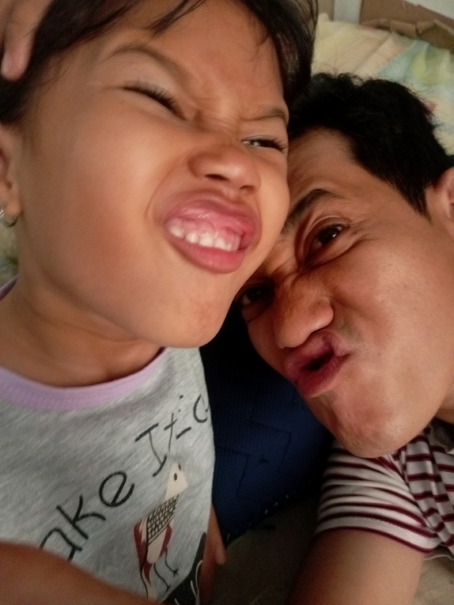 Two individuals, a child and an adult male, are positioned closely together, both making funny faces for a selfie. The child, on the left, has their eyes squinted shut and tongue sticking out. The adult male, on the right, is also squinting and puckering their lips. The child is wearing a grey t-shirt with a graphic of a bird and text that appears to read "ake It's..." . The adult male is wearing a striped shirt. The background suggests an indoor setting, possibly a home in Batam, Indonesia, with a patterned fabric visible behind them. The overall mood is playful and intimate.