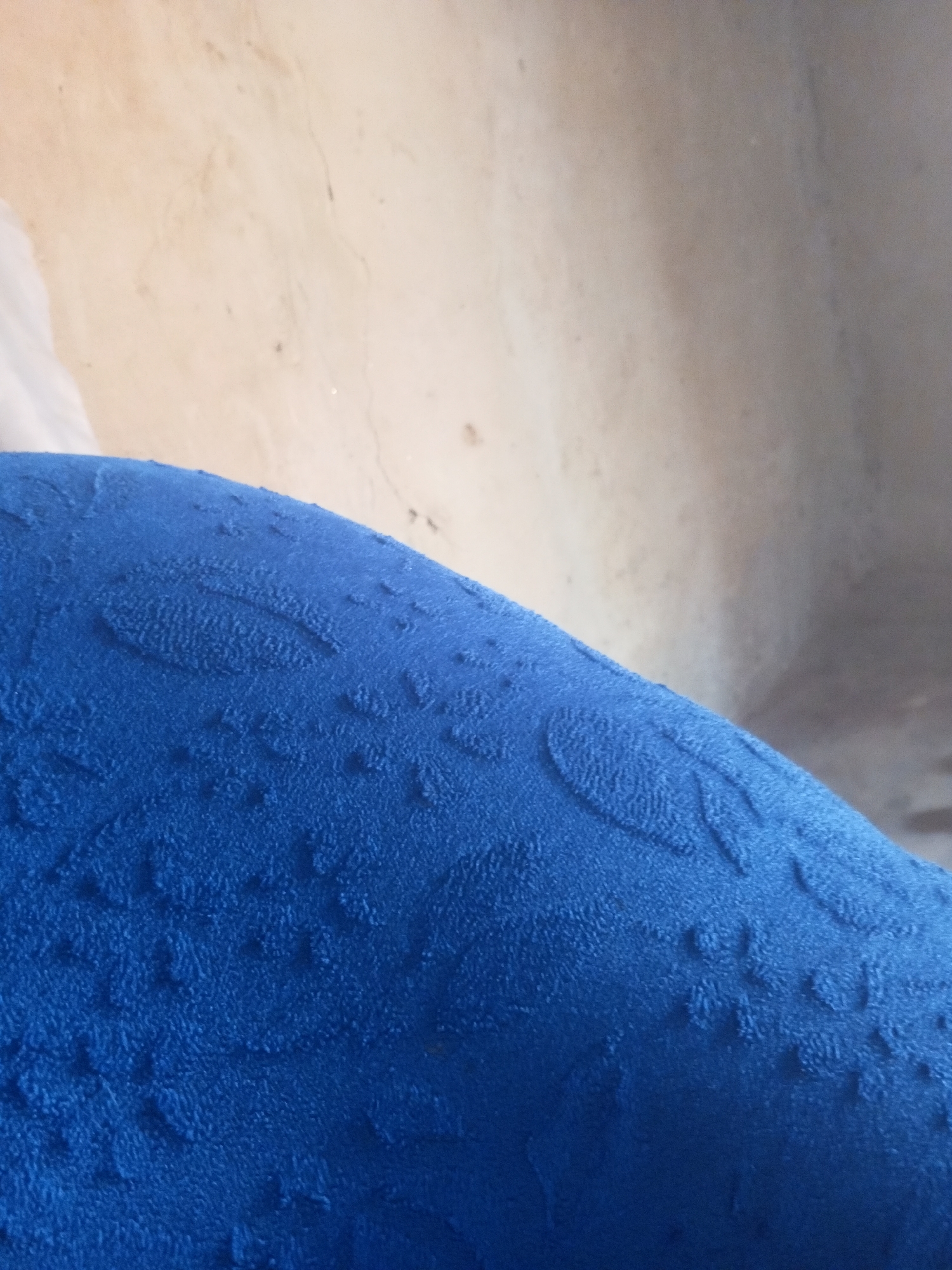 The image shows a close-up of a textured blue fabric, possibly a cushion or upholstery. The fabric has a raised, looped pattern that appears to be consistent across its surface. In the background, a wall with a textured, off-white surface is visible, with some subtle cracking or imperfections. The lighting suggests an indoor setting, though no specific urban features are discernible. The image does not provide any information about the city of Nyabe, Nigeria, nor does it depict any people, actions, or interactions. It is a purely still-life composition focused on the texture and color of the fabric and the background wall.
