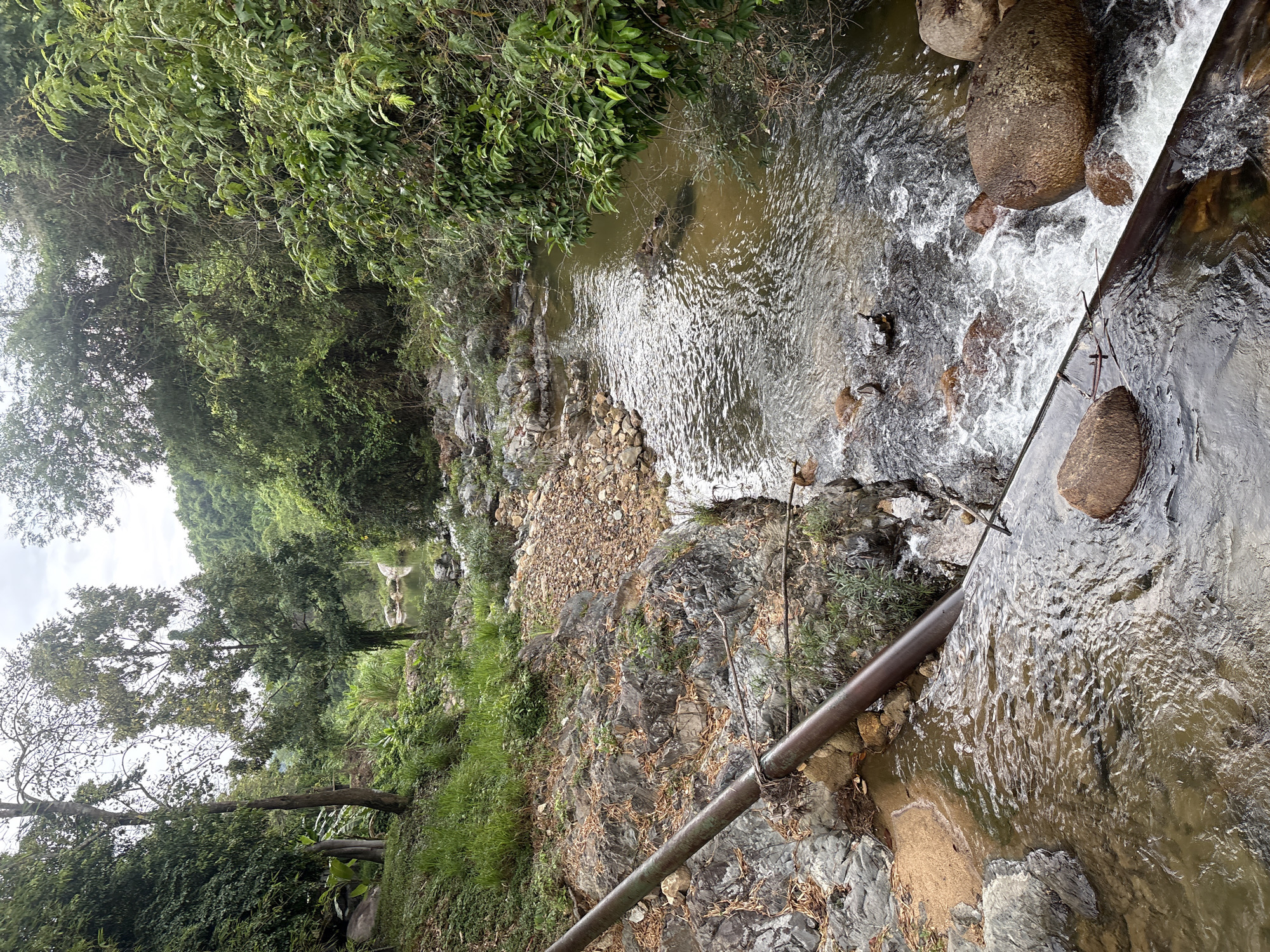 This image depicts a stream flowing through a rocky, forested area. The location is implied to be in Khanh Vinh, Vietnam. The stream has sections of rapid flow and areas where the water appears calmer. Boulders are present within the stream. Vegetation, including trees and bushes, lines the banks. A pipe runs along a section of the stream's edge. No people are visible in the image.