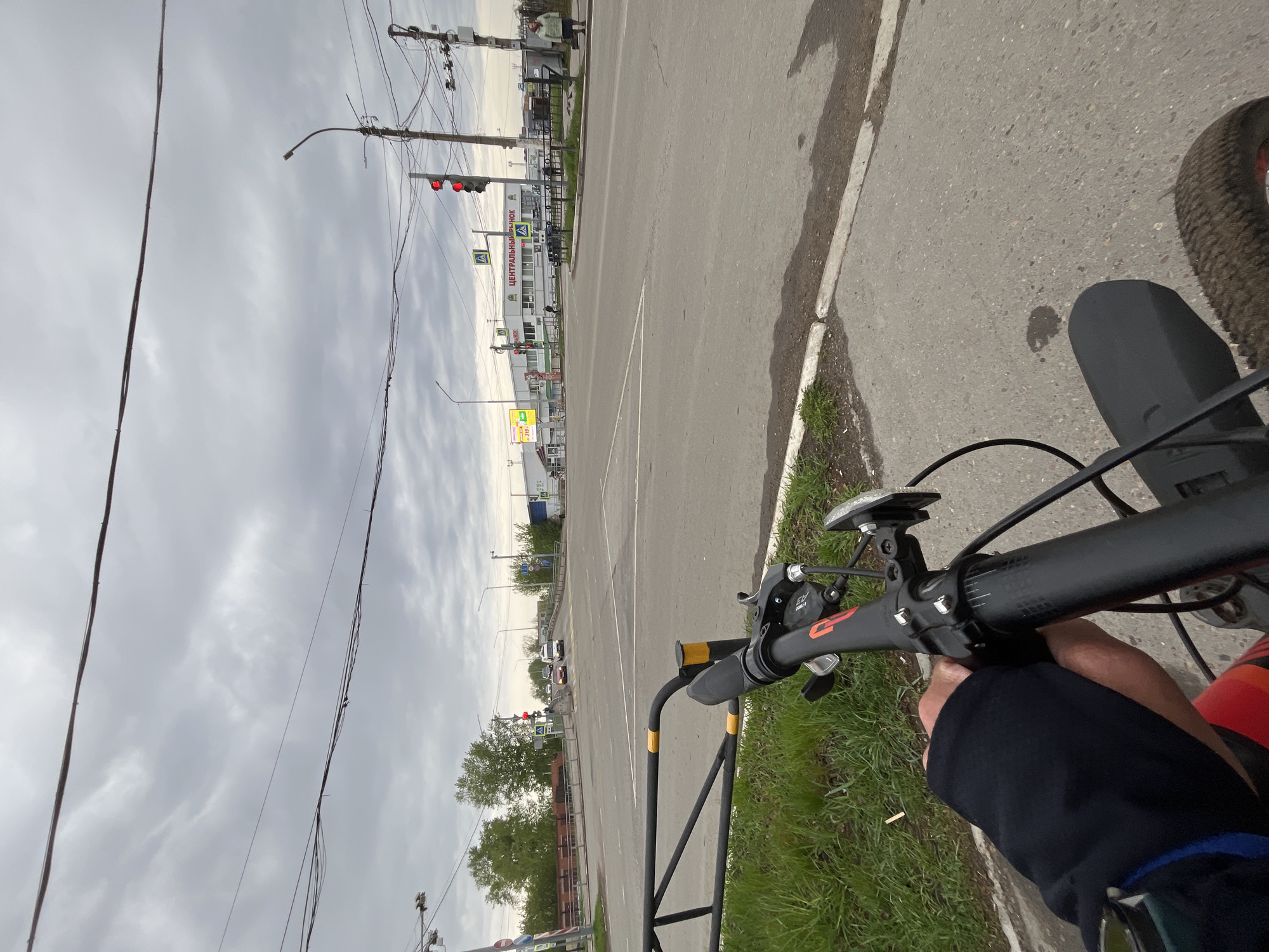 View from a bicycle stopped at a traffic light on Sovetskaya Street in Chaikovsky, Russia. Red traffic light visible. Sign for "Central Market" (Центральный рынок) in Russian. Cloudy sky. Black bicycle handlebars with lights and controls visible. A person's leg clad in dark clothing is visible. A road and sidewalk extend into the distance. Grass grows alongside the road. A black barrier is near the bike.