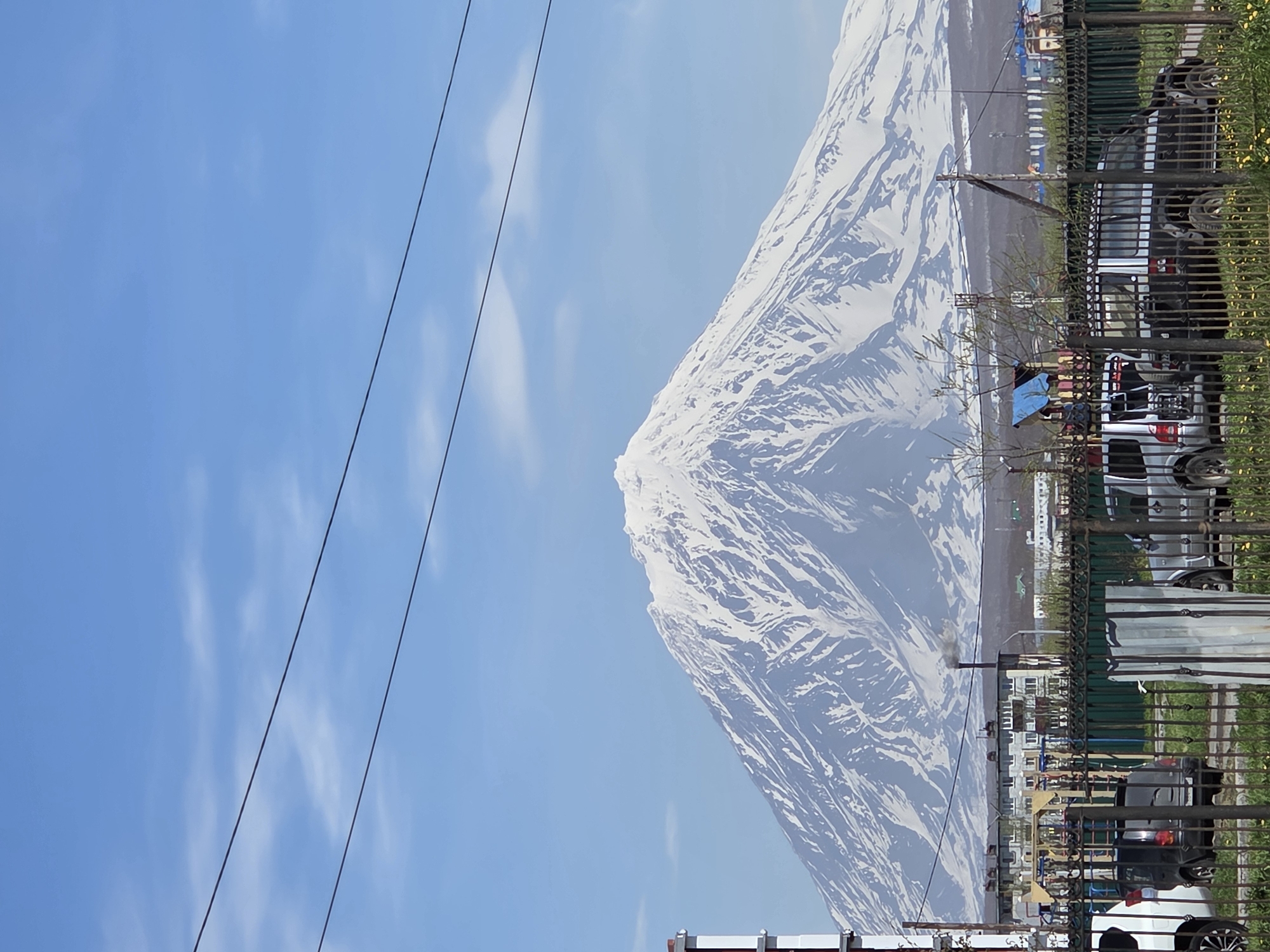 The image displays a clear daytime scene in the city of Unknown, Unknown, under a light blue sky with scattered faint white clouds. Two dark, parallel power lines stretch horizontally across the upper portion of the frame. A large, snow-capped stratovolcano dominates the background, its peak and upper slopes covered in white snow with visible dark rock formations. Below the mountain, the mid-ground reveals a residential area with several multi-story apartment buildings, typically featuring light-colored facades and dark roofs. One building exhibits a chimney emitting a plume of dark grey smoke. A colorful children's playground structure with blue, yellow, and red elements is partially visible behind a fence. The foreground is dominated by a black metal fence with decorative finials, behind which are parked multiple vehicles: a partially visible white SUV on the left, a dark hatchback, a dark SUV with a rear-mounted bicycle rack, and another dark SUV on the far right. The ground in the foreground is covered in green grass interspersed with yellow dandelions. Wooden utility poles are visible throughout the mid-ground. No individuals are present in the depicted scene.