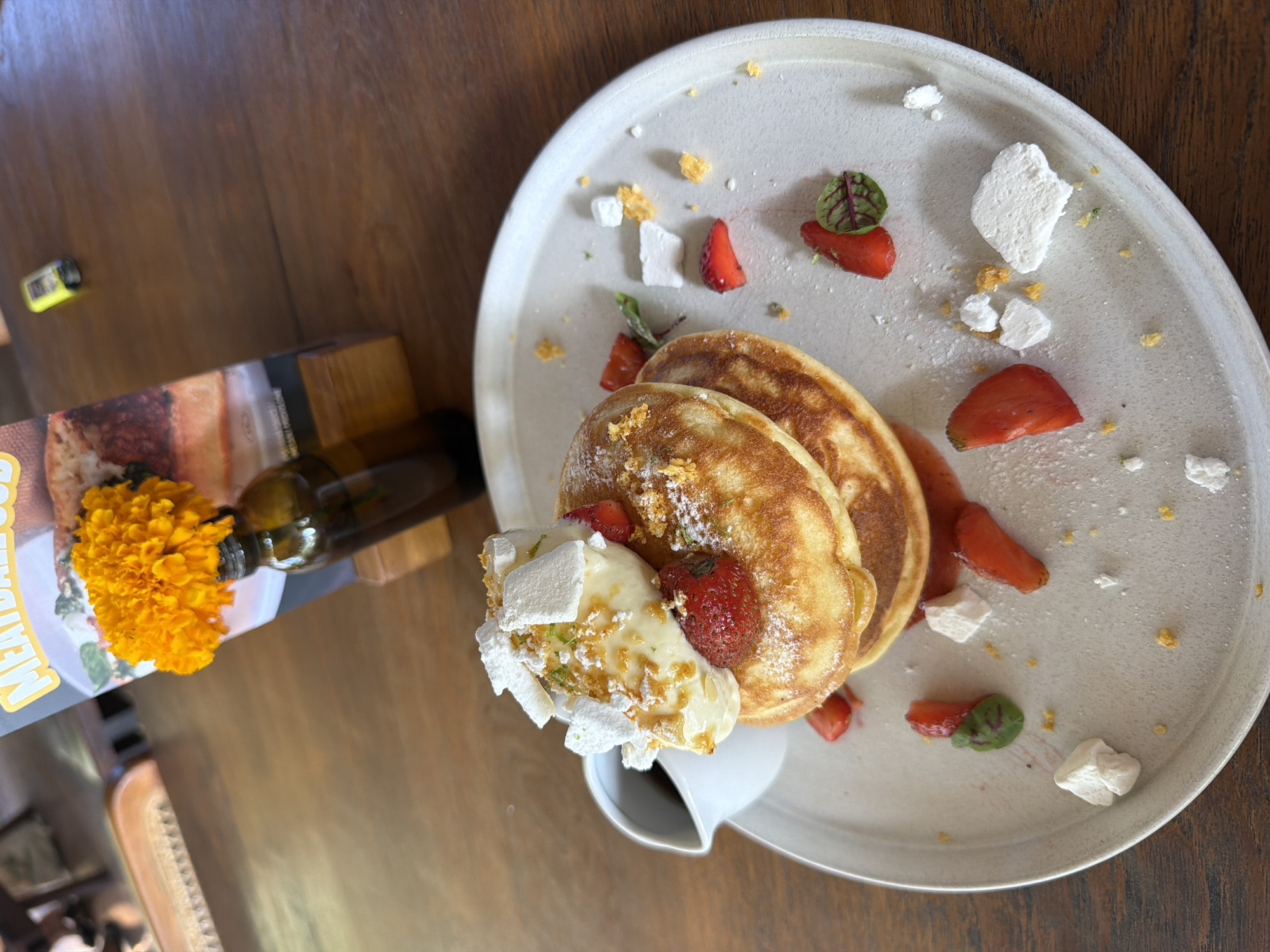 A close-up, eye-level shot captures a dessert plate on a wooden table in Kuta Selatan, Indonesia. The plate holds a stack of three golden-brown pancakes, generously topped with a dollop of creamy white topping, a scattering of bright red strawberry slices, and delicate white meringue pieces. Powdered sugar dusts the pancakes and the surrounding plate. A small white pitcher, containing a dark liquid, sits to the left of the pancakes. Scattered on the plate are additional pieces of strawberries, meringue, and small yellow crumbs. In the background, out of focus, is a menu with an orange flower in a dark glass bottle. A small yellow object, possibly a lighter, rests on the table further back. The lighting suggests it is daytime.