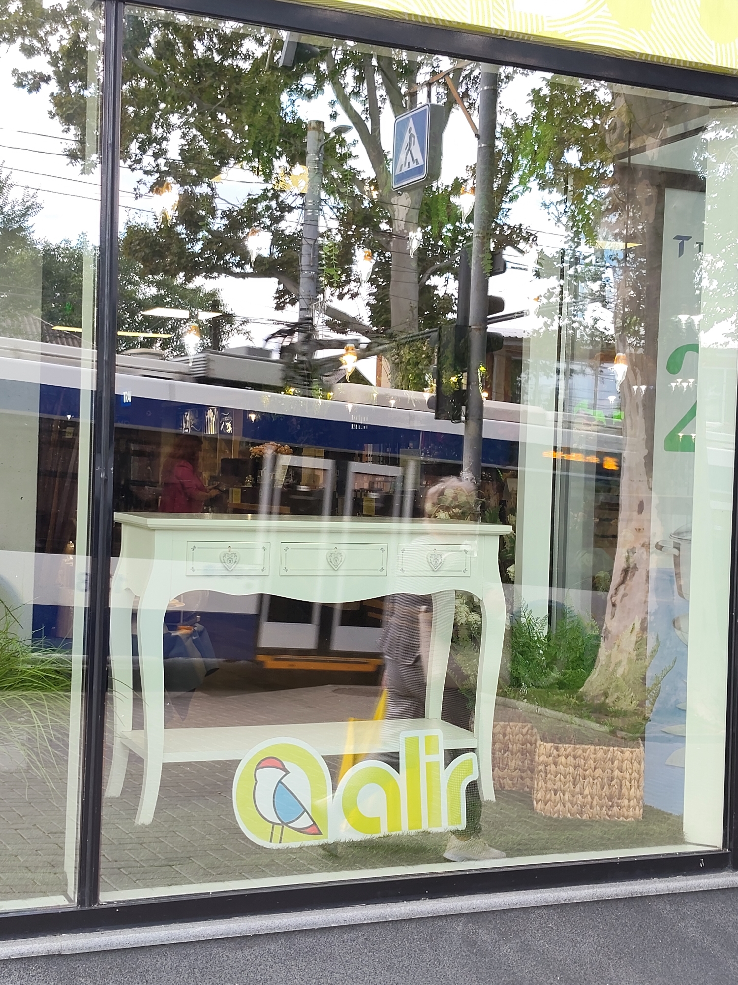 The image shows a street scene reflected in a shop window in Chișinău, Moldova. The reflection displays a blue and white tram passing by, with several people visible inside. In front of the shop window, a light green console table with decorative drawers is displayed. A logo featuring a stylized bird and the word "alr" in yellow is also visible on the window. The background reflection includes trees, power lines, and a pedestrian crossing sign, indicating an urban environment. The lower part of the window shows the street pavement.