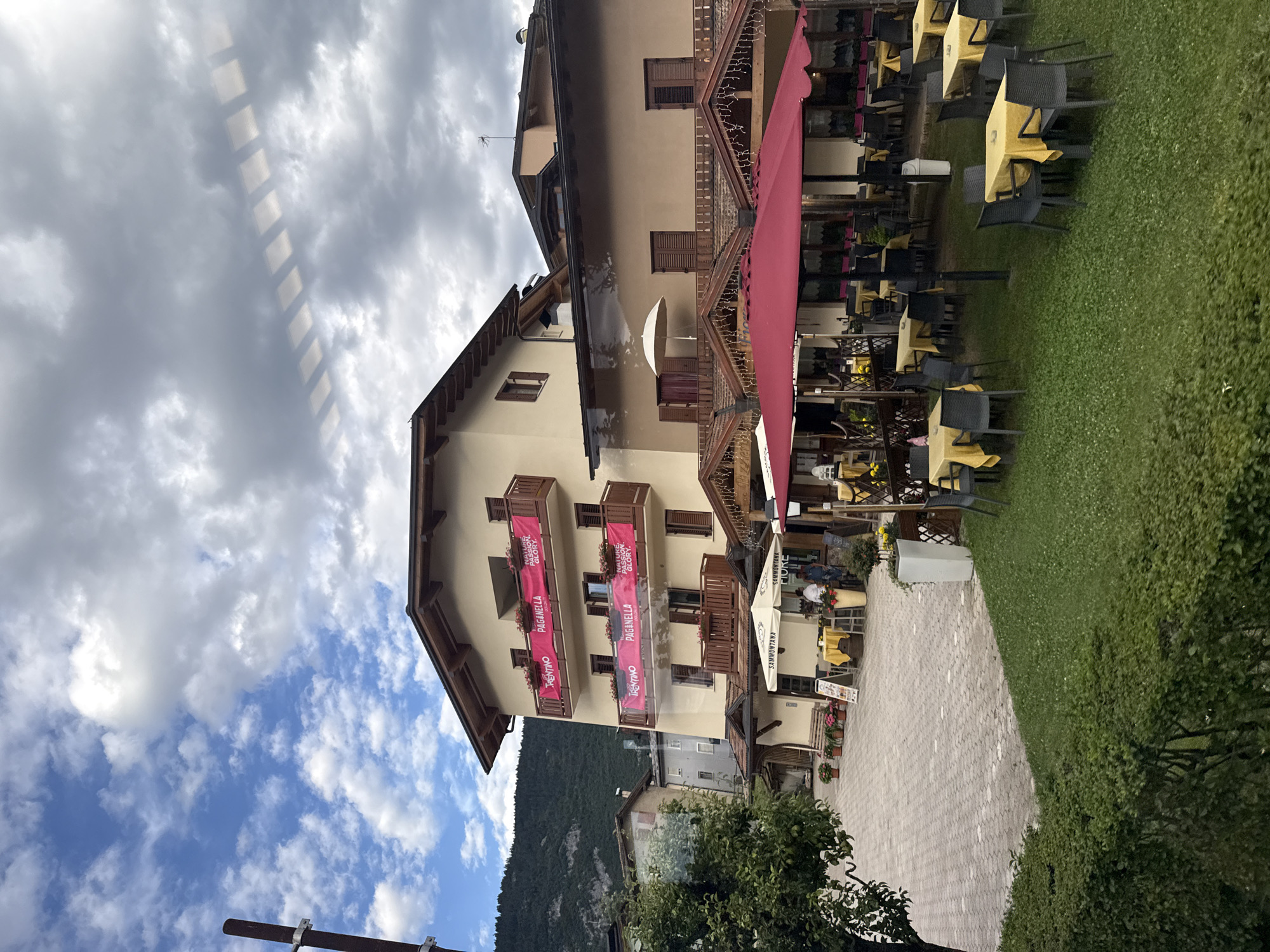 The image shows a street scene in Cavedago, Italy, featuring a building with banners and an outdoor dining area. The building is multi-storied with a cream-colored facade and brown shutters. Pink banners with the text "TRENTINO" and "PAGANELLA" are displayed on balconies. An outdoor seating area is set up in front of the building, with numerous tables covered in yellow tablecloths and surrounded by chairs. Some tables have umbrellas providing shade. In the background, a hillside with trees is visible, and the sky is partly cloudy. The ground in front of the building is paved with interlocking bricks, and a grassy area with more tables and chairs is also present. A few people are seen near the tables, and one person appears to be working at a counter.