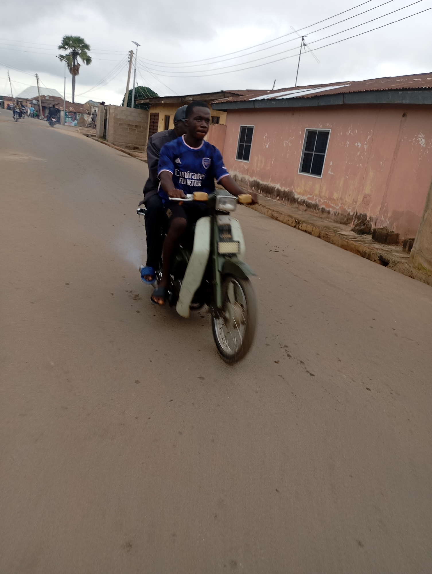 Two young males are riding on a motorcycle on a paved road in Shendam, Nigeria. The rider in front is wearing a blue and white soccer jersey with the text "Emirates Fly Better" and black pants, with blue and black sandals on his feet. The passenger behind him is wearing a dark jacket. The motorcycle is green and white and appears to be older. The road is a light brown color and has some scattered dark spots. On the right side of the road, there is a long, pale pink building with two dark windows and a corrugated metal roof. The background shows a cloudy sky, power lines, utility poles, a palm tree, and other buildings. The scene appears to be in a rural or semi-urban area.
