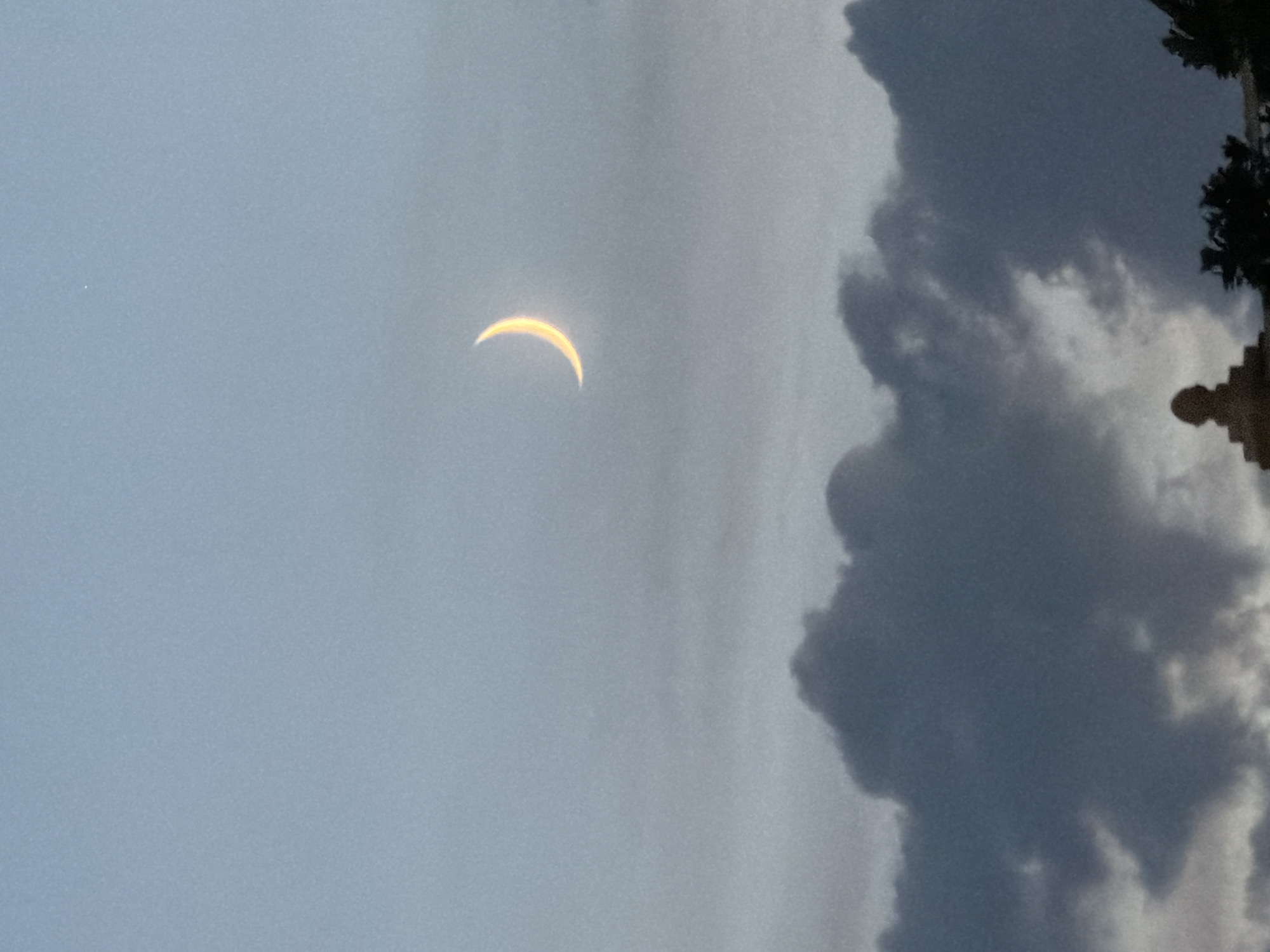 A crescent moon hangs in a hazy, pale blue sky. Dark, billowy clouds fill the lower third of the image, with the silhouette of a building or monument and some trees visible at the very bottom.