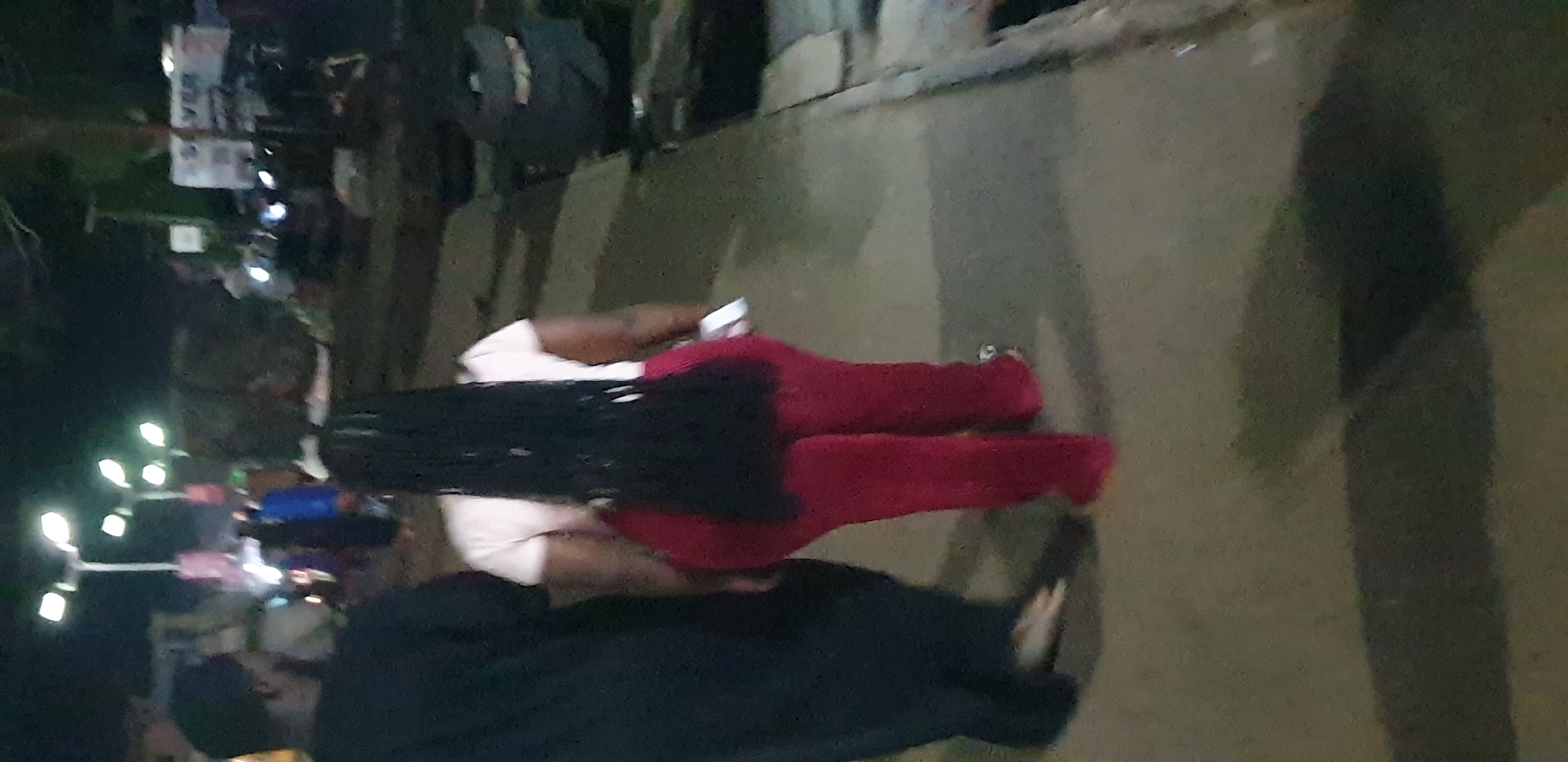 The image captures a nighttime street scene in Sabon Tasha, Nigeria. Two individuals are walking away from the camera on a paved pathway. The person on the left wears a dark, loose-fitting garment, with their bare feet or light-colored footwear partially visible. The person on the right wears a light-colored short-sleeved top and dark red pants. Their long, dark braided hair extends down their back. This individual is holding a small, light-colored object in their right hand. The ground is a dark, uneven paved surface. A raised curb or edge is visible along the right side of the pathway, beyond which dark, indistinct objects, possibly refuse or equipment, are present. In the blurry mid-ground, multiple streetlights with bright, multi-faceted lamps illuminate the area. Further in the background, numerous indistinct human figures are visible among blurred architectural structures, suggesting a populated commercial or residential street. A partially visible sign in the upper right background displays the word "SILVER".