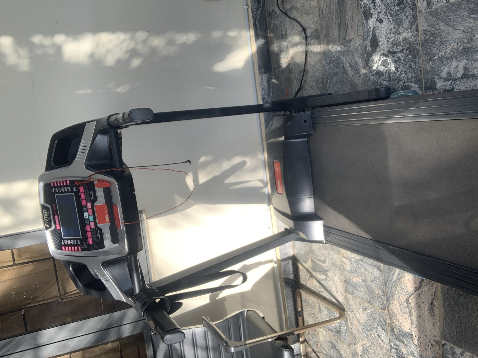 A Fytter RU-06R treadmill is positioned on a speckled grey tiled floor. The treadmill is black and silver, with a digital display screen at the front. Red and black wires are visible connected to the display panel. A black chair with a metallic frame is located to the left of the treadmill. The scene is illuminated by sunlight casting shadows on a white wall in the background. The location is identified as Abuja, Nigeria.
