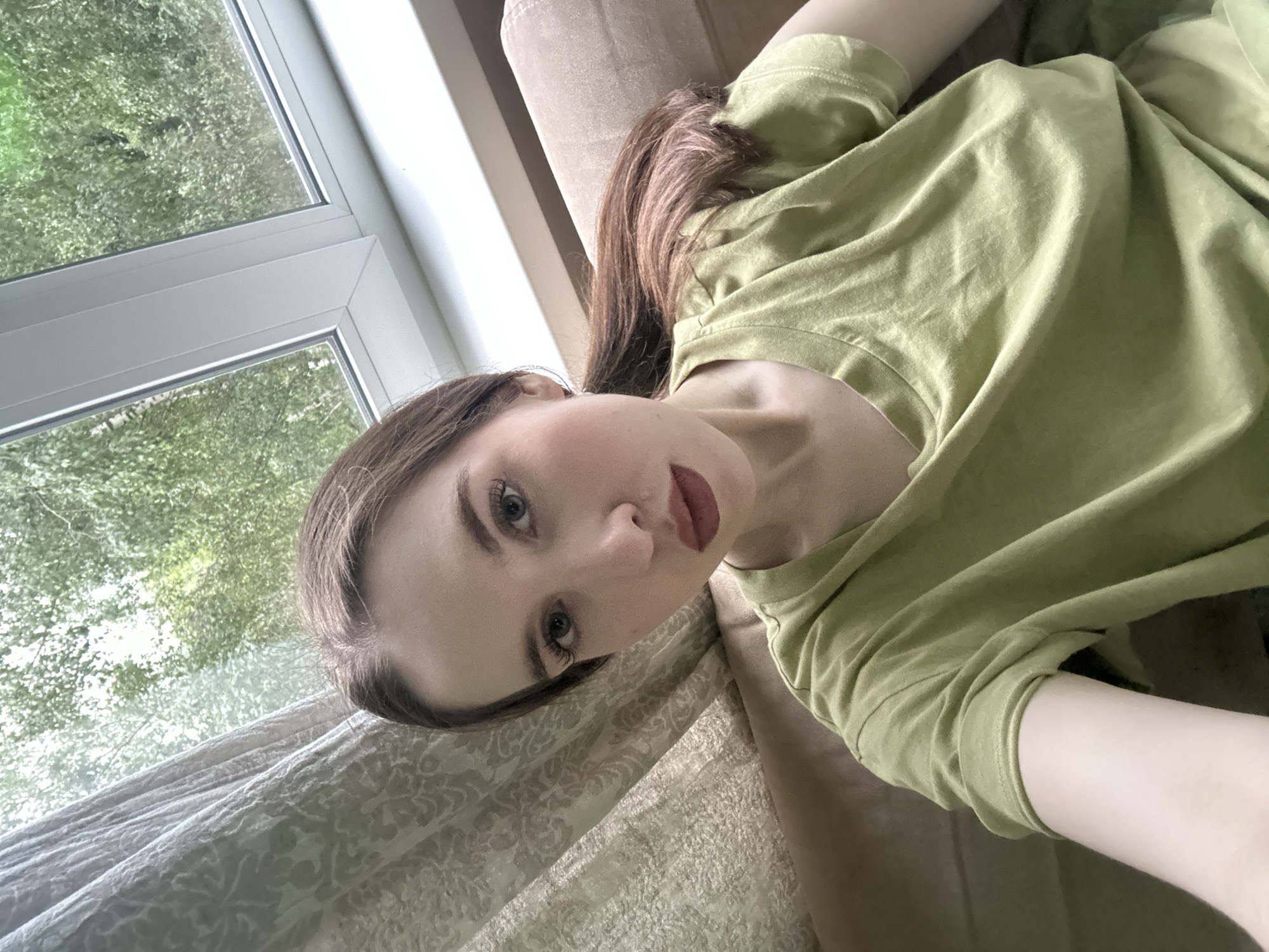 A young woman with light brown hair tied in a ponytail, wearing a light green t-shirt, is taking a selfie indoors. She is seated on a couch in front of a window that looks out onto lush green trees. A patterned curtain hangs to the left of the window. The scene is indoors, suggesting a residential setting, possibly in Moscow, Russia, given the foliage visible through the window. The woman's makeup is subtle, with emphasis on her eyes and lips.