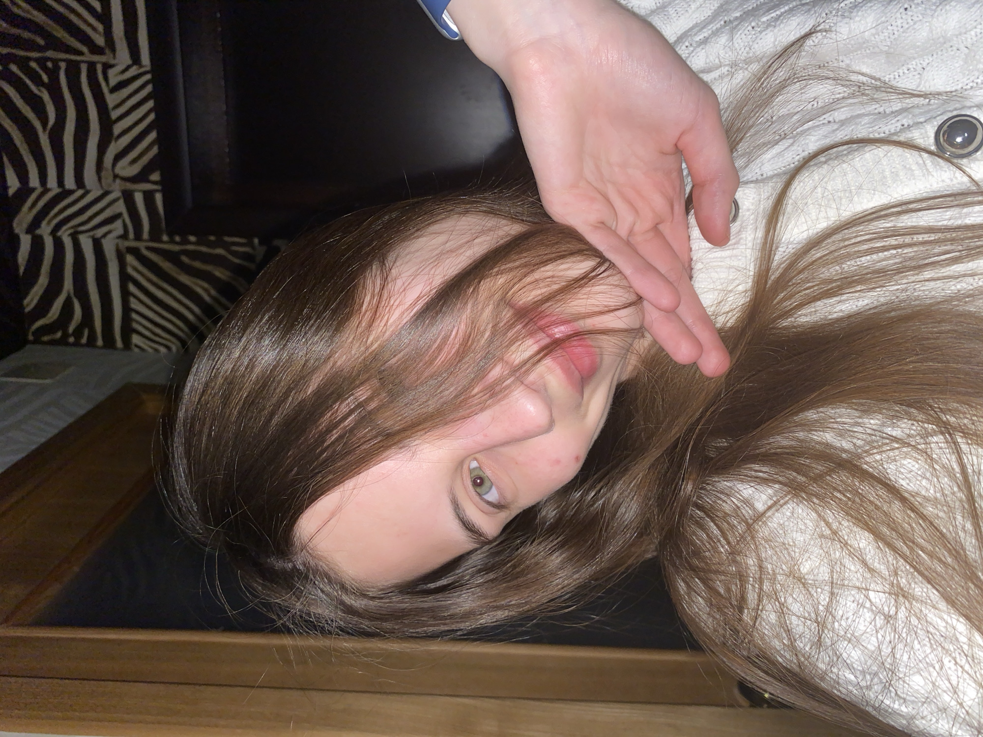 An overhead, close-up shot captures a young person, likely female, with long, straight brown hair and greenish-hazel eyes, lying down and looking directly at the viewer. The image is rotated 90 degrees clockwise, positioning the subject's head towards the bottom of the frame. Their right hand is gently placed near their face, with fingers partially obscuring their mouth and chin. A blue strap, possibly from a watch or bracelet, is visible on their right wrist.
The person is wearing a white, textured garment, resembling a knitted sweater, which is spread out around them. Two prominent round, silver-toned buttons are visible on this clothing. The long brown hair cascades around their head and across parts of the white garment. Minor red marks or blemishes are visible on the nose and cheek area.
The background features a distinctive black and off-white zebra-stripe pattern to the upper left, likely part of furniture upholstery or wall decor. Below the subject's head, a dark, smooth surface gives way to a wooden border at the very bottom of the frame. The lighting suggests an indoor setting. No text is visible in the image.