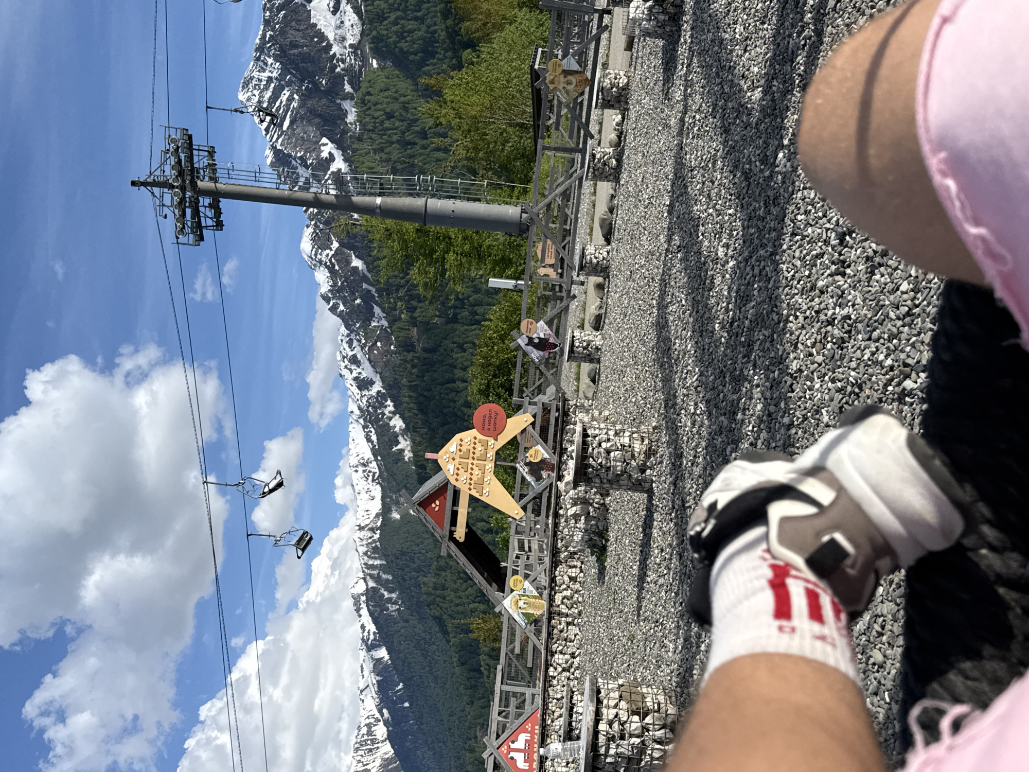 The image presents a vibrant, sunlit outdoor scene captured from a first-person perspective at a mountain resort, likely in Russia. In the immediate foreground, the viewer's legs and feet are visible. The person wears white athletic socks with prominent red stripes and a logo, paired with beige, white, and black sneakers. A pink garment, possibly shorts or a shirt, is also seen. The ground beneath is covered in small, light gray gravel.
The midground features a well-maintained viewing or recreational area. A sturdy wooden fence with gabion-style bases (wire cages filled with rocks) extends across the frame. Along this fence, various interpretive signs and decorative wooden structures are displayed. A prominent feature is a large, stylized wooden cut-out representing a village or architectural complex, topped with a red circular sign that reads in Cyrillic: "МАМА, Я ЗДЕСЬ ЖИВУ!" (MAMA, I LIVE HERE!). Another sign closer to the foreground displays "РОДНИК" (SPRING). A child is visible near these displays, seemingly observing them. A large metal ski lift tower stands tall, supporting several cables that stretch across the landscape.
The background reveals a spectacular natural panorama. Majestic snow-capped mountains dominate the horizon, their lower slopes covered in dense, dark green coniferous forests. The sky is a brilliant blue with scattered fluffy white clouds. Empty ski lift chairs are seen suspended from the cables, traversing the clear sky.
The scene suggests a leisurely moment at a high-altitude tourist destination, possibly a ski resort operating during warmer months. The bright daylight and clear weather indicate excellent conditions for outdoor enjoyment. The overall impression is one of natural beauty combined with recreational facilities.