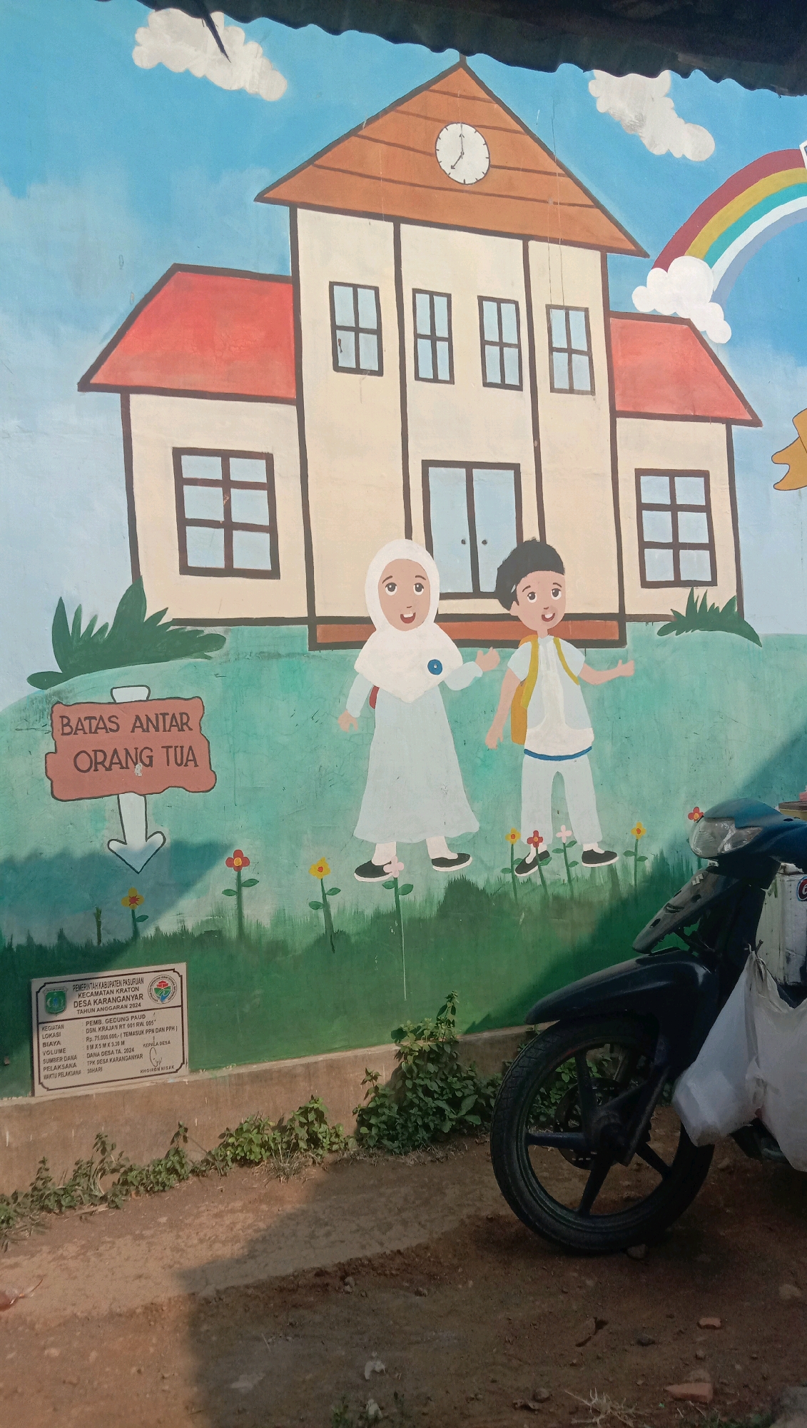 A vibrant mural on a wall depicts a two-story building with a clock on its roof, a rainbow arching in the sky, and two children. The left child, a girl wearing a hijab and a light blue dress, is gesturing outwards. The right child, a boy in a white shirt, yellow vest, and blue pants, also has his arms extended. A sign to the left of the children reads "BATAS ANTAR ORANG TUA" (Parent Drop-off Limit) with an arrow pointing down. At the bottom left of the mural is a plaque with text, which appears to be official information for a PAUD (early childhood education) building located in the village of Karanganyar, Karanganyar Regency, Indonesia, with a budget of Rp. 75,500,000 for construction in 2024. In the foreground, a black motorcycle is parked, with a white plastic bag hanging from its handlebars. The ground is a mix of dirt and concrete, with some overgrown vegetation around the base of the wall. The scene appears to be outdoors, under daylight.