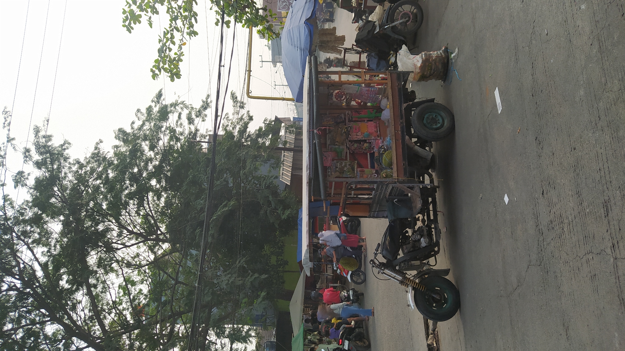 In Klapanunggal, Indonesia, a three-wheeled vehicle, modified with a cargo bed that functions as a mobile stall, is parked on an asphalt road. The stall displays various items, including what appear to be durians and colorful merchandise. The vehicle is positioned in front of other market stalls, some covered by tarps, suggesting a roadside market environment. Several people are visible in the background, some near motorcycles, and others are interacting at the market stalls. The scene is characterized by an overcast sky, large trees providing partial shade, and overhead electrical wires, indicative of an urban or semi-urban setting. The overall atmosphere suggests a typical day at a local marketplace.