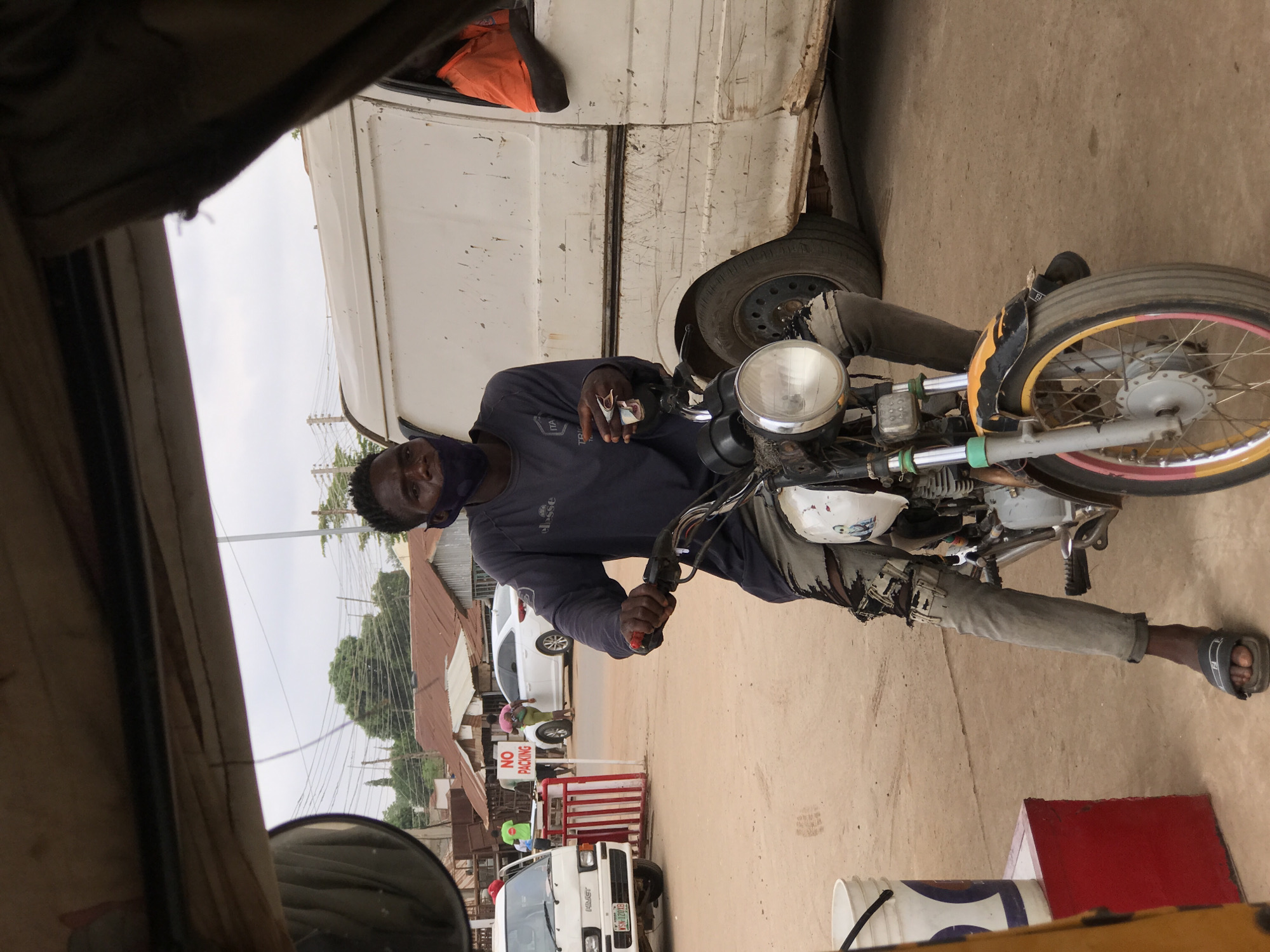 A man wearing a face mask and a dark blue long-sleeved shirt is sitting on a motorcycle. He is holding money in one hand. He is wearing faded grey trousers and sandals. The motorcycle is parked next to a white van. The front tire of the motorcycle has a multi-colored rim. A "NO PARKING" sign is visible in the background. The scene appears to be a street in Abuja, Nigeria. Other vehicles and buildings are visible in the background. The photo is taken from inside another vehicle, possibly a taxi or auto-rickshaw.