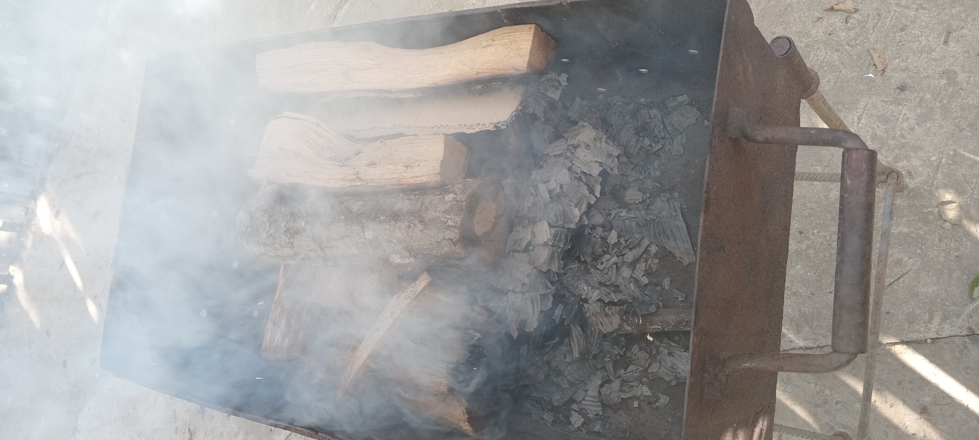 A top-down view of a rusty metal barbecue grill filled with partially burned logs and ashes. Smoke billows up from the grill, obscuring some of the contents. The grill is positioned outdoors on a concrete surface, likely in the city of Koviahy, Ukraine, as indicated by the context. The surrounding environment appears to be a casual outdoor setting, possibly a backyard or a park. The lighting suggests it is daytime, with shadows visible on the ground. There are no people or animals present in the image.