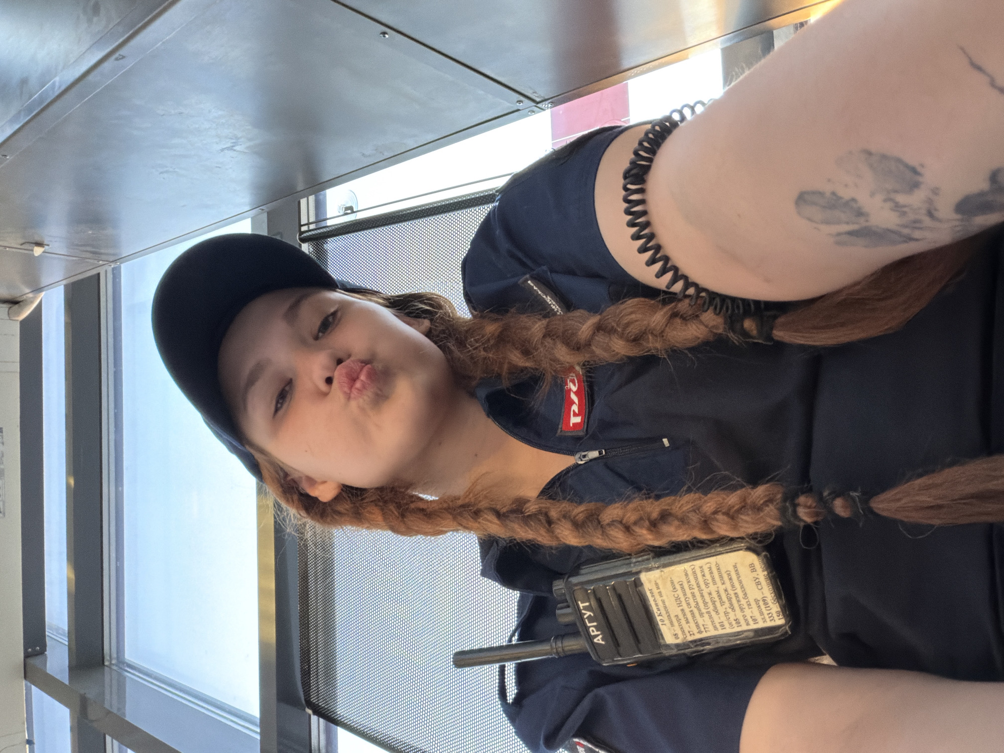 This media file shows a close-up, low-angle selfie of a young woman with brown hair in braids, wearing a dark blue uniform shirt, a black baseball cap, and a coiled black bracelet. She is making a kissing face at the camera. A black radio device is clipped to her shirt. The background suggests an indoor setting with large windows offering a view of a bright sky, indicating daytime. The woman has a tattoo on her upper arm. The text on the radio device appears to be in Russian, listing various codes and phrases, possibly for emergency services or security personnel. One visible patch on her shirt has the word "Pro" on it. Based on the location context provided, the setting could be in Rostov-on-Don, Russia. The overall mood of the image is playful and casual.